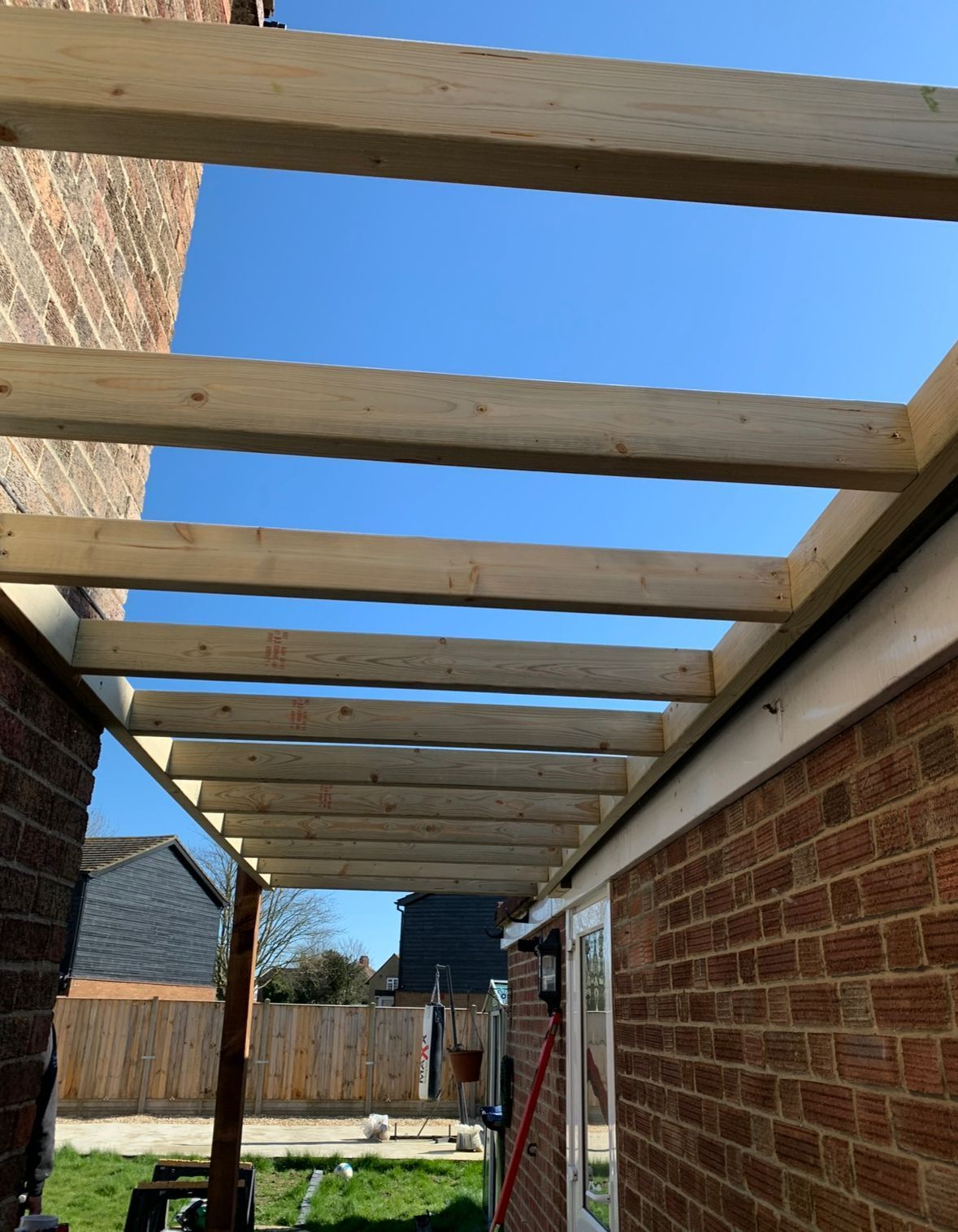 Wooden pergola under construction, attached to a brick house, against a blue sky.
