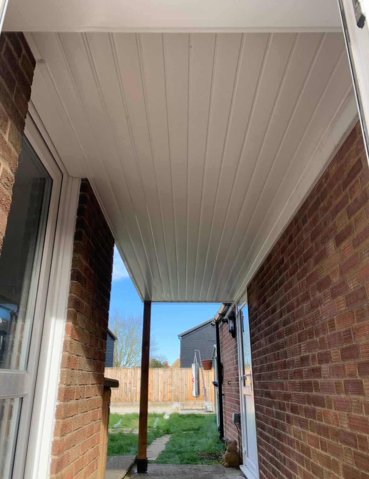 White paneled ceiling over a porch supported by a wooden post, flanked by brick walls and a view of a backyard.