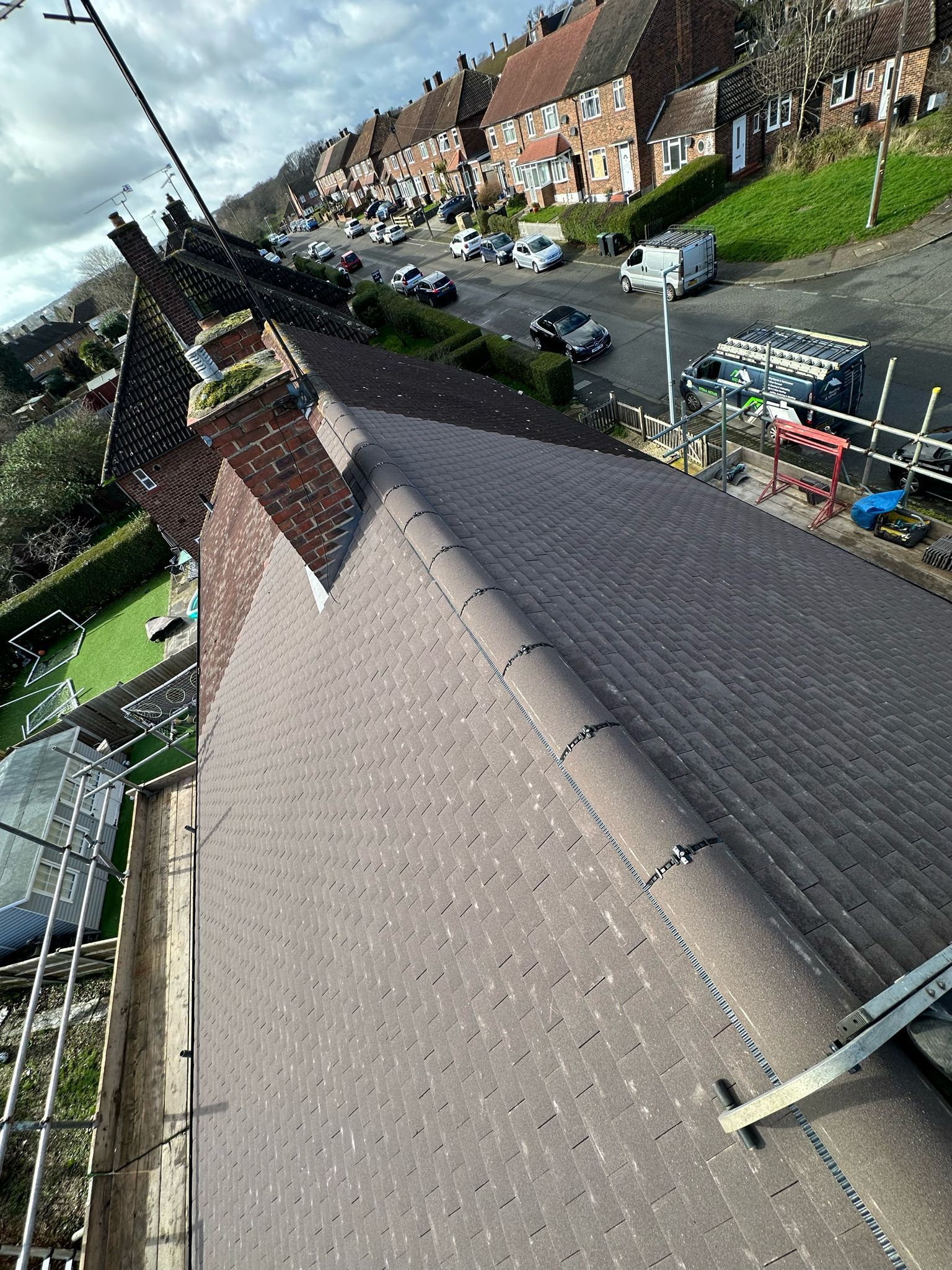 Rooftop view of a residential street. Brown roof, green grass, parked cars, and houses on a hill.