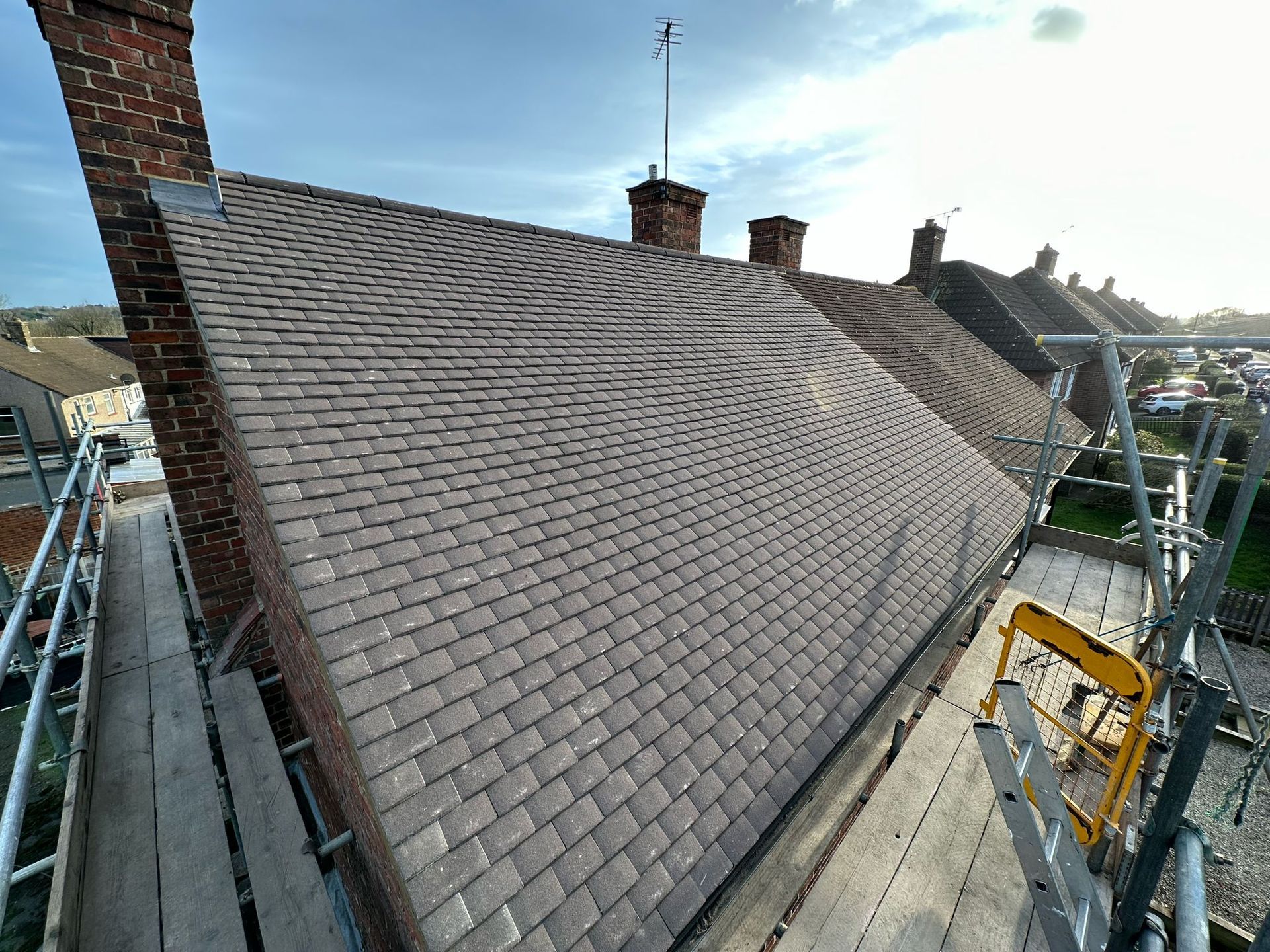 Roof with dark gray tiles, scaffolding, brick chimney, antenna, and overcast sky.