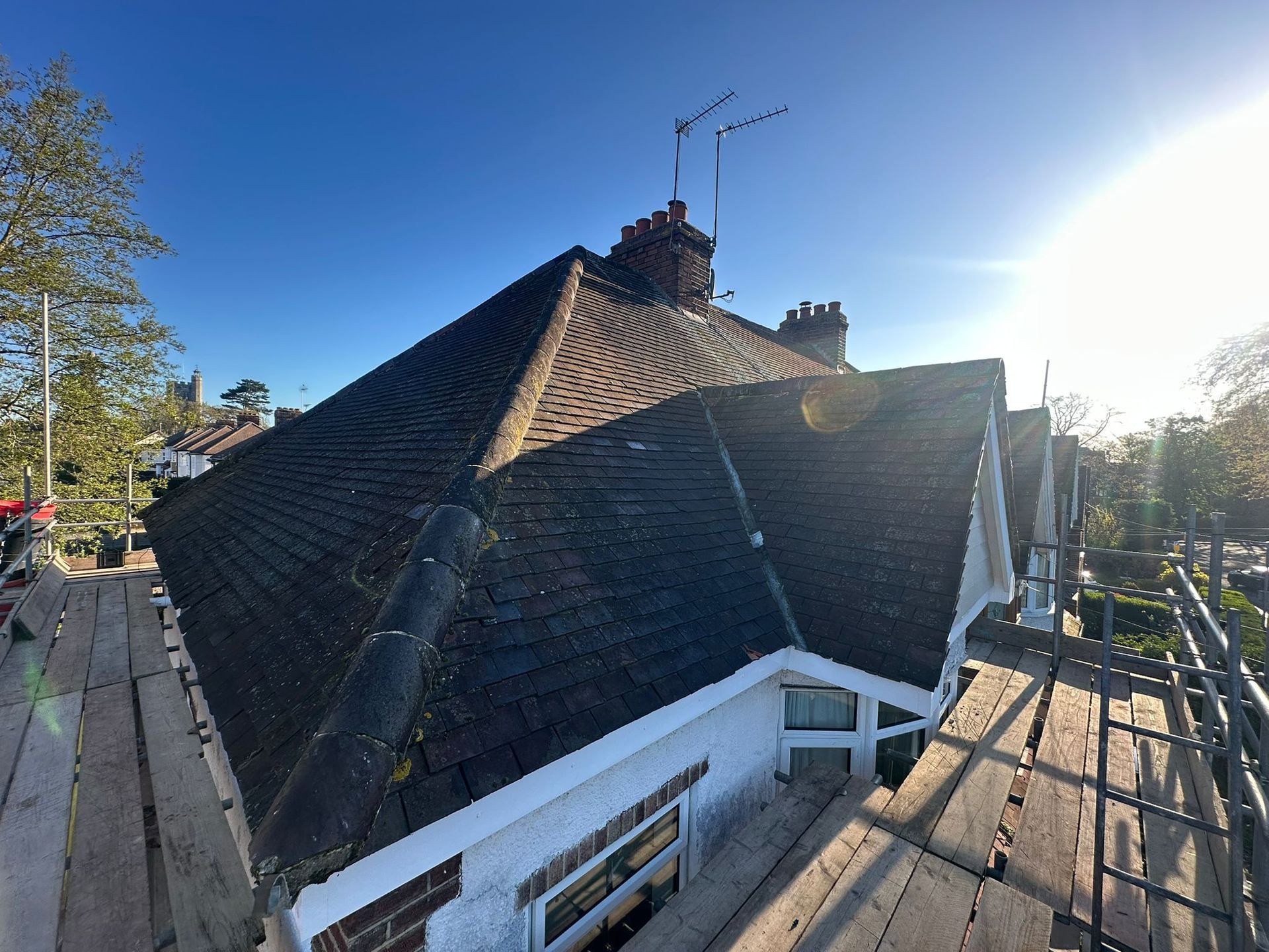 Steep shingled roof with chimney and bright sun, viewed from a wooden deck with trees nearby