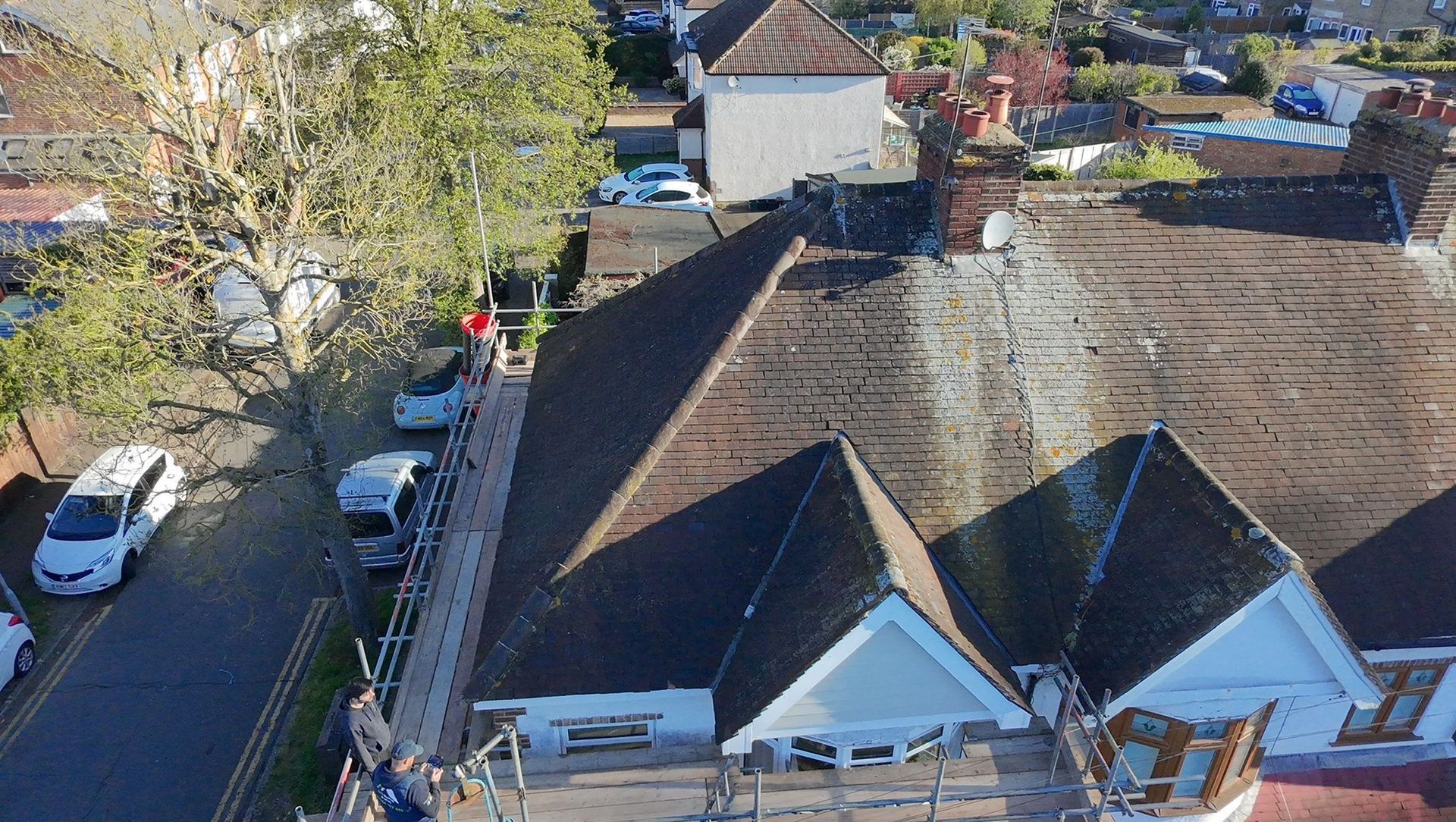 Aerial view of a residential street with parked cars, trees, and rooftops in daylight.