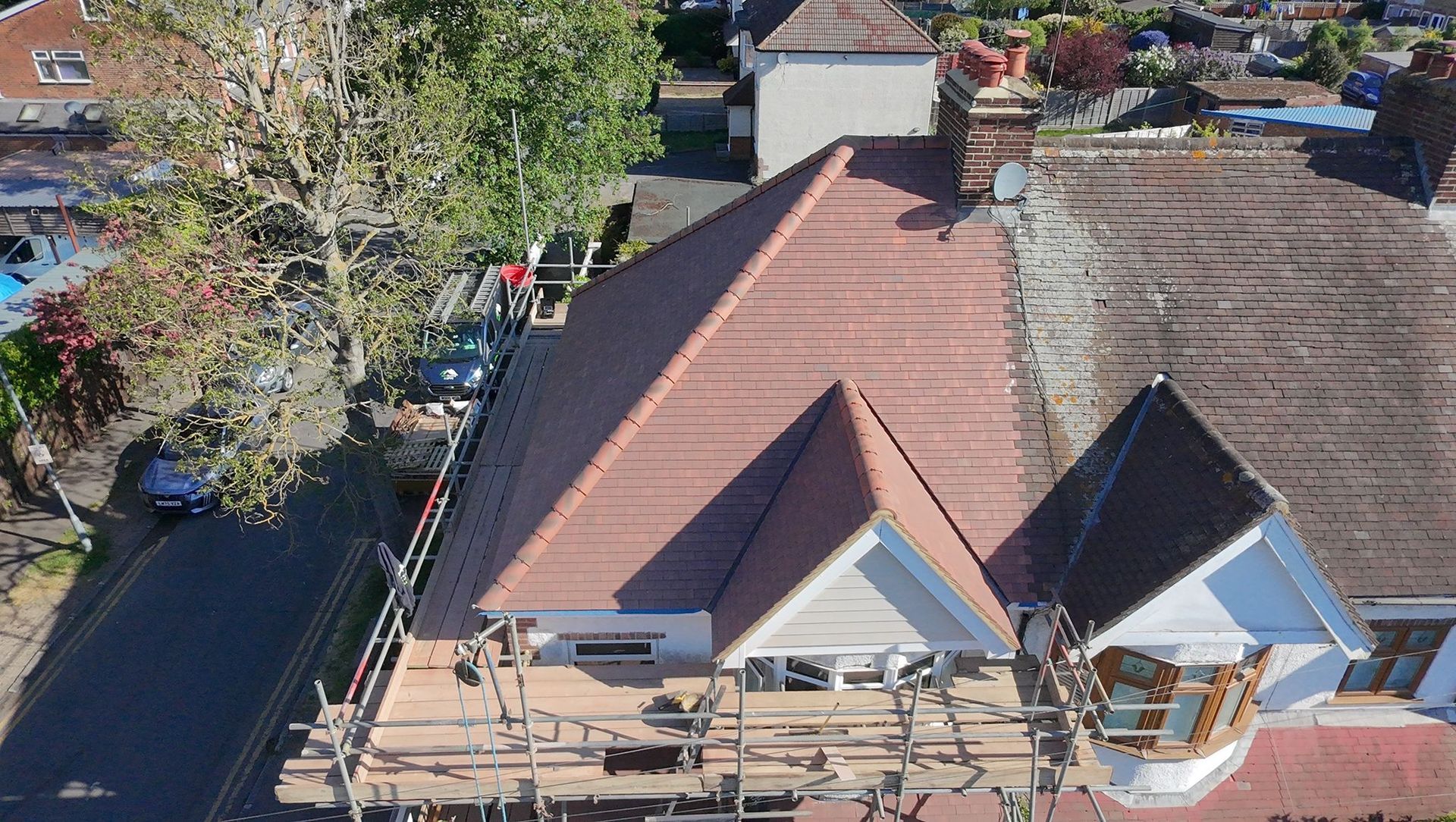 Aerial view of a house with a red roof, dormer windows, and a patio bordered by trees and a street.