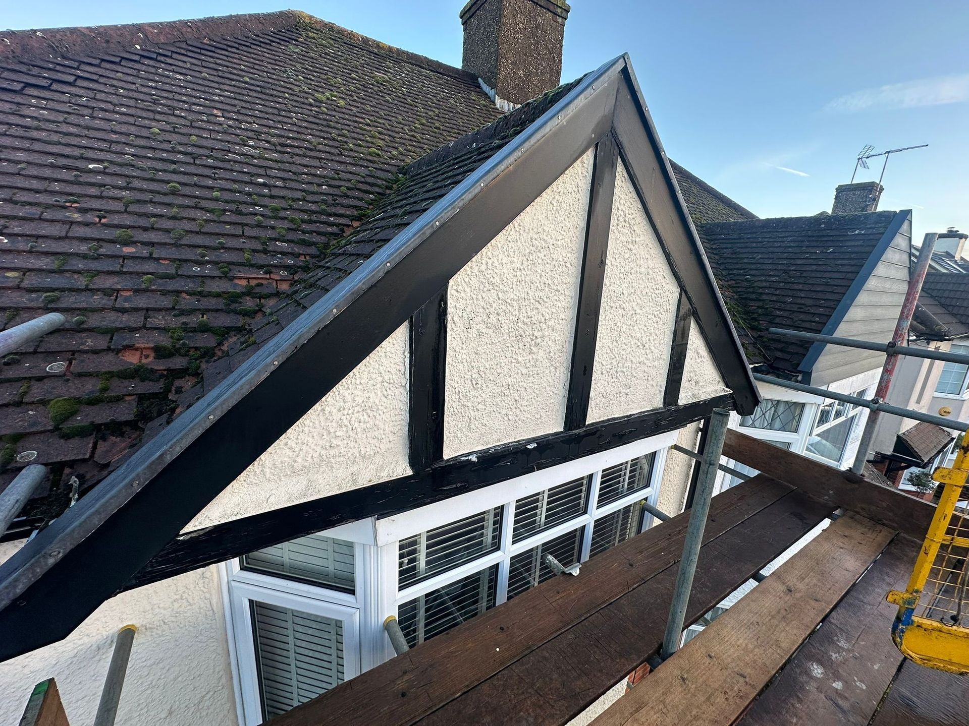 Black and white trim on a house gable, with a scaffolding setup. Dark roof tiles, blue sky.