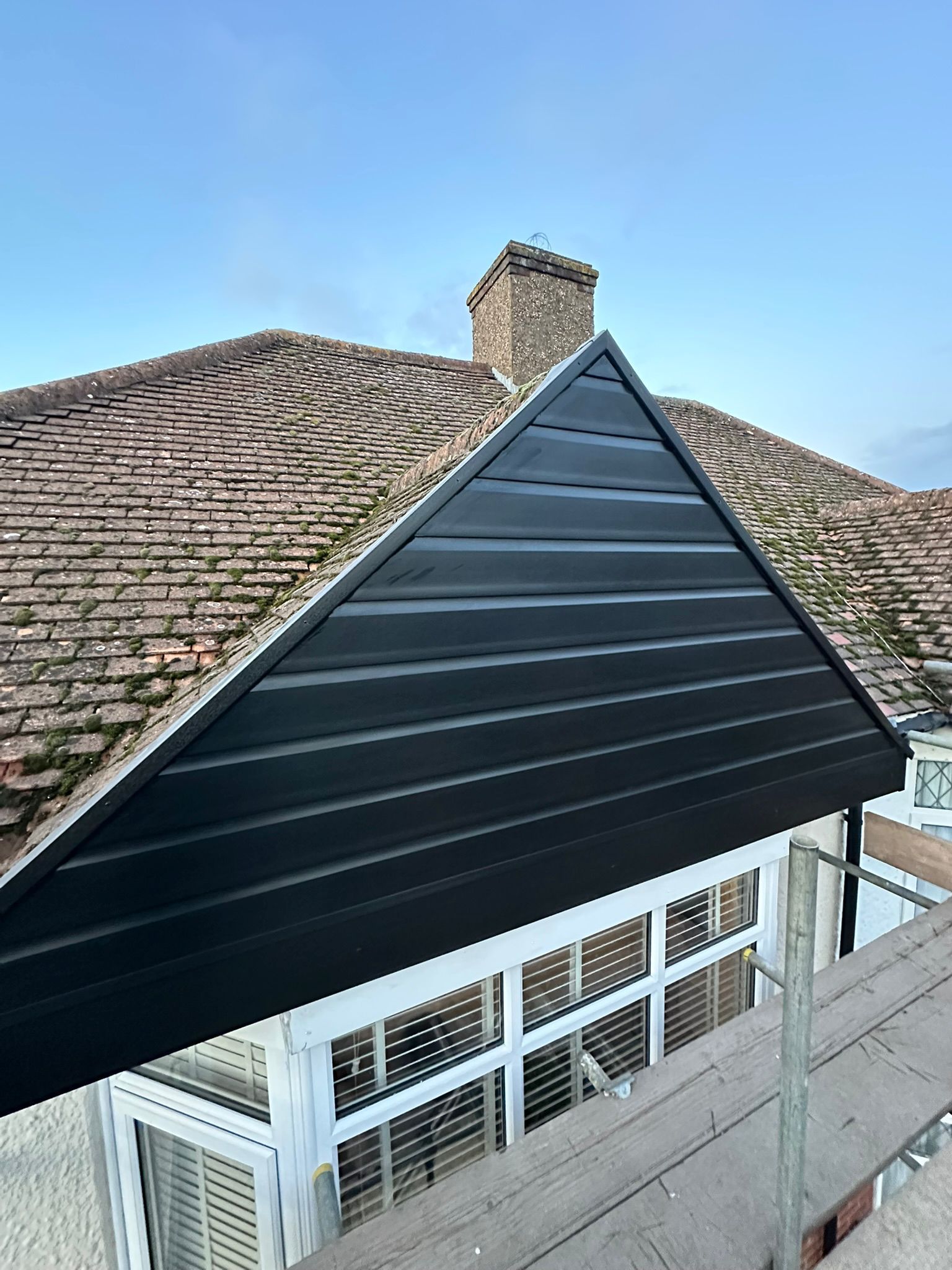 Black metal roof section on a building with weathered tile roof and chimney under a blue sky.