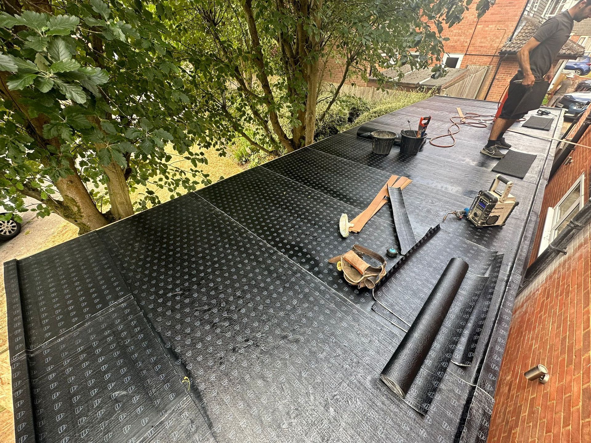 Roofer working on a flat black roof with tools. Trees and a brick building are in the background.