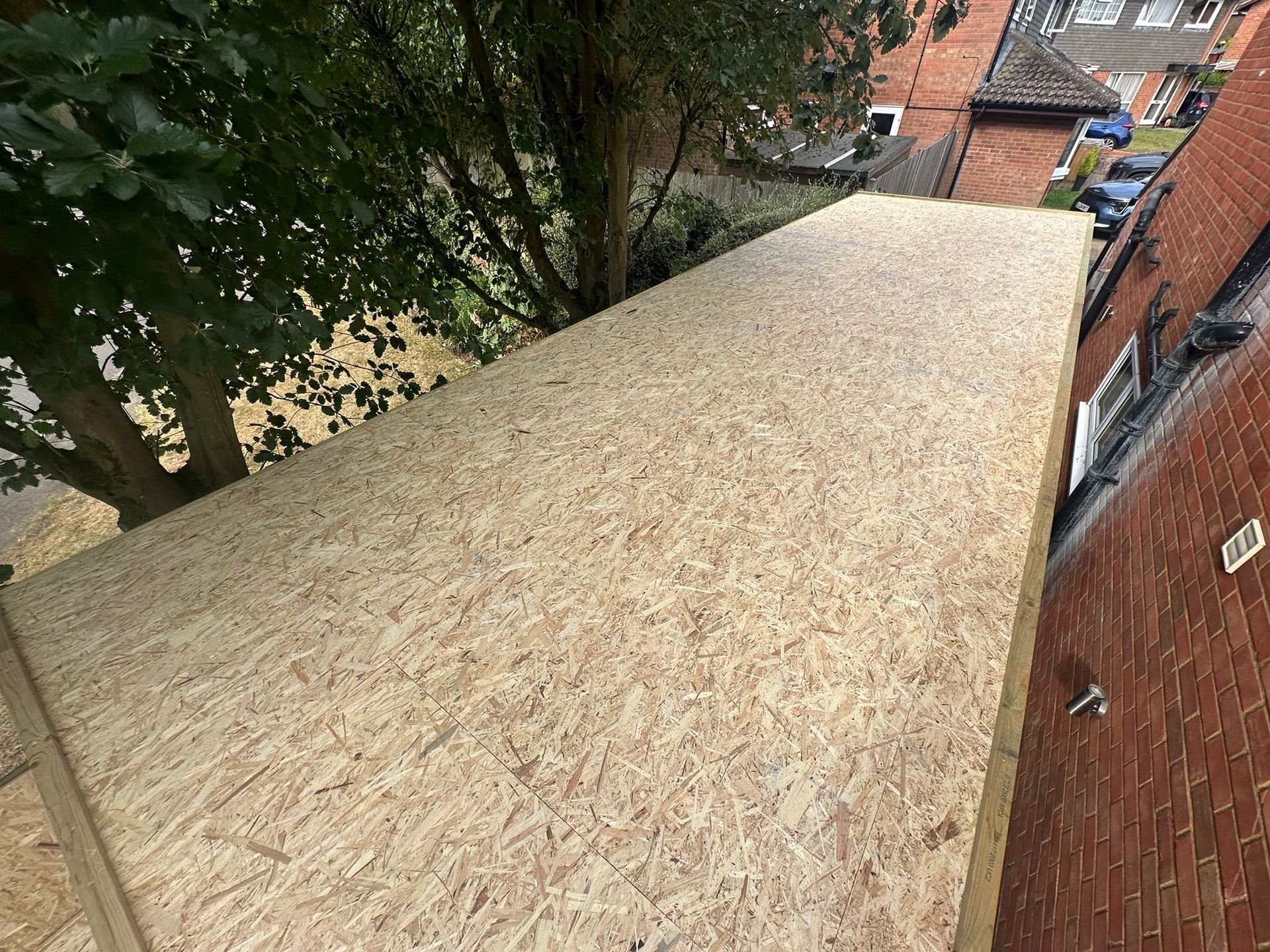 OSB-sheathed roof extension. Tan, textured wood. Viewed from above, near a tree and brick building.