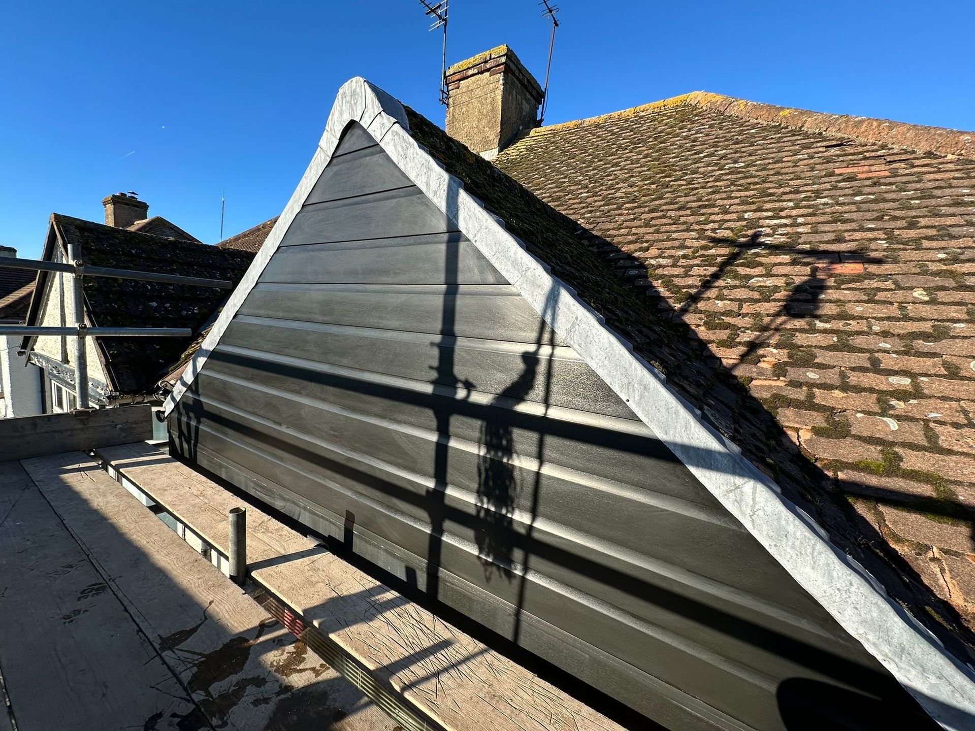A triangular black siding gable on a roof, with scaffolding below and an adjacent tiled roof.