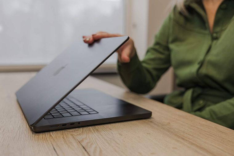 Iemand opent een donkere laptop op een licht houten bureau, binnenshuis.