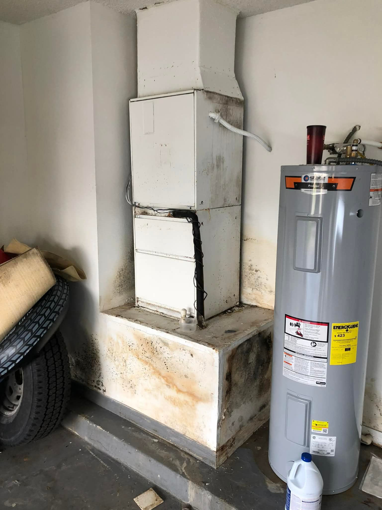 Water heater and vent pipe in a garage corner beside a gray tank, with a tire and debris nearby.
