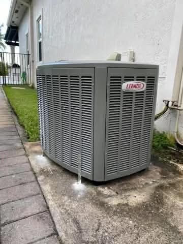 Outdoor gray air conditioner unit beside a house wall on a wet concrete pad with gravel nearby