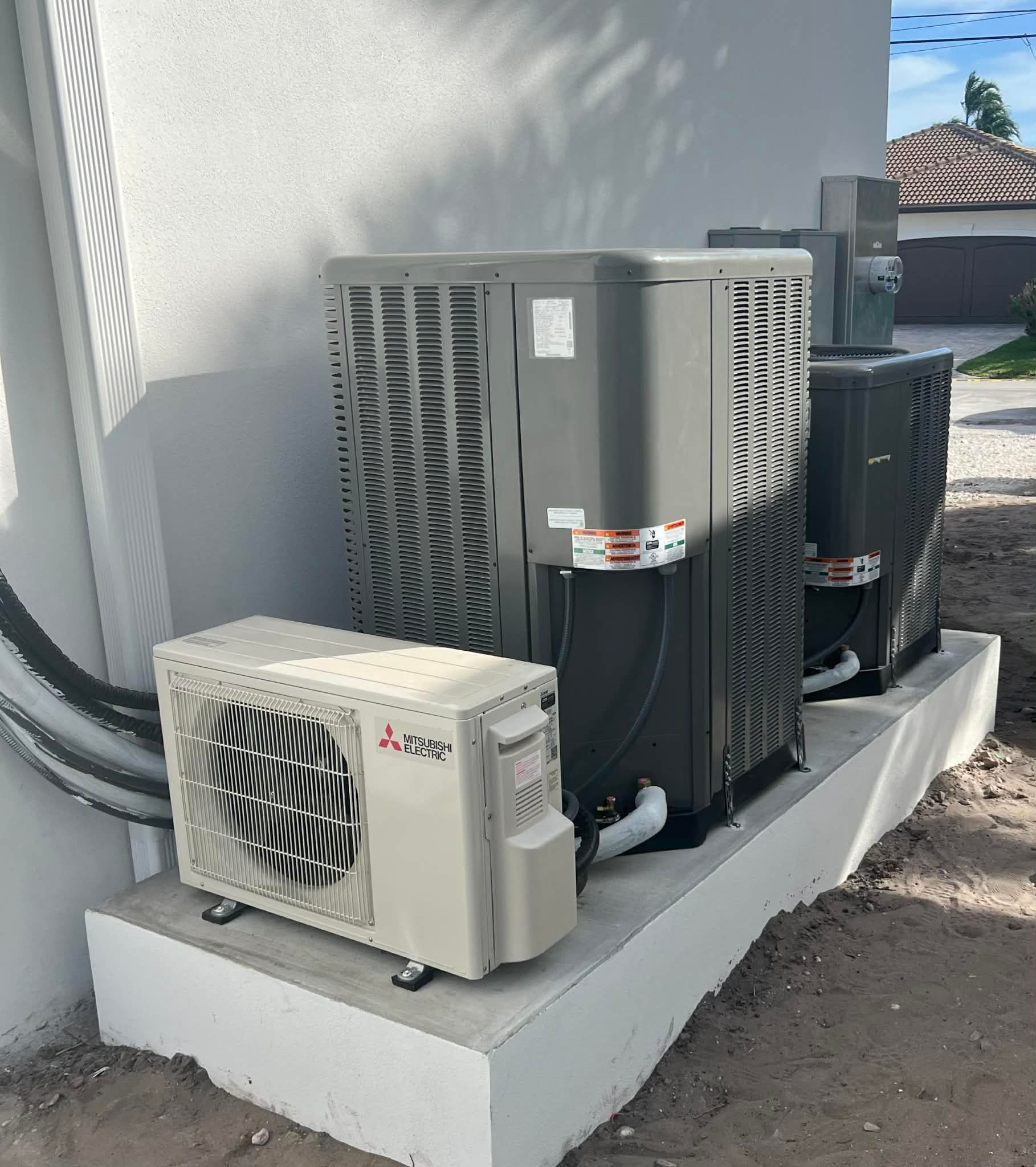 Three outdoor HVAC units beside a house on a concrete pad, including a beige mini-split condenser and two gray units