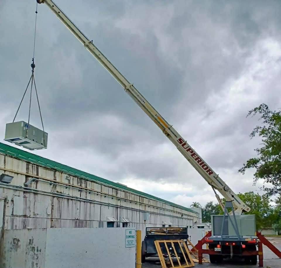 Crane lifting a gray equipment box beside a building under cloudy skies