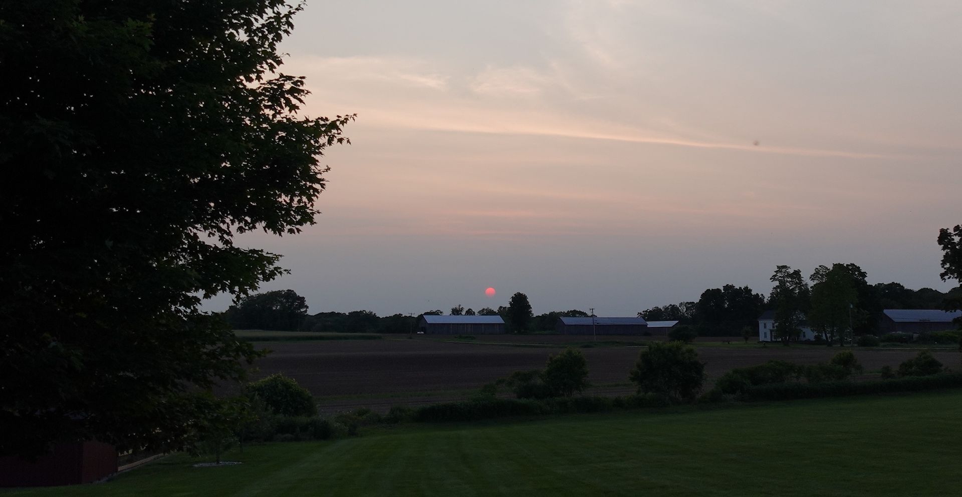 Sunset over a rural landscape with fields, trees, and buildings; sky is pink and gray.