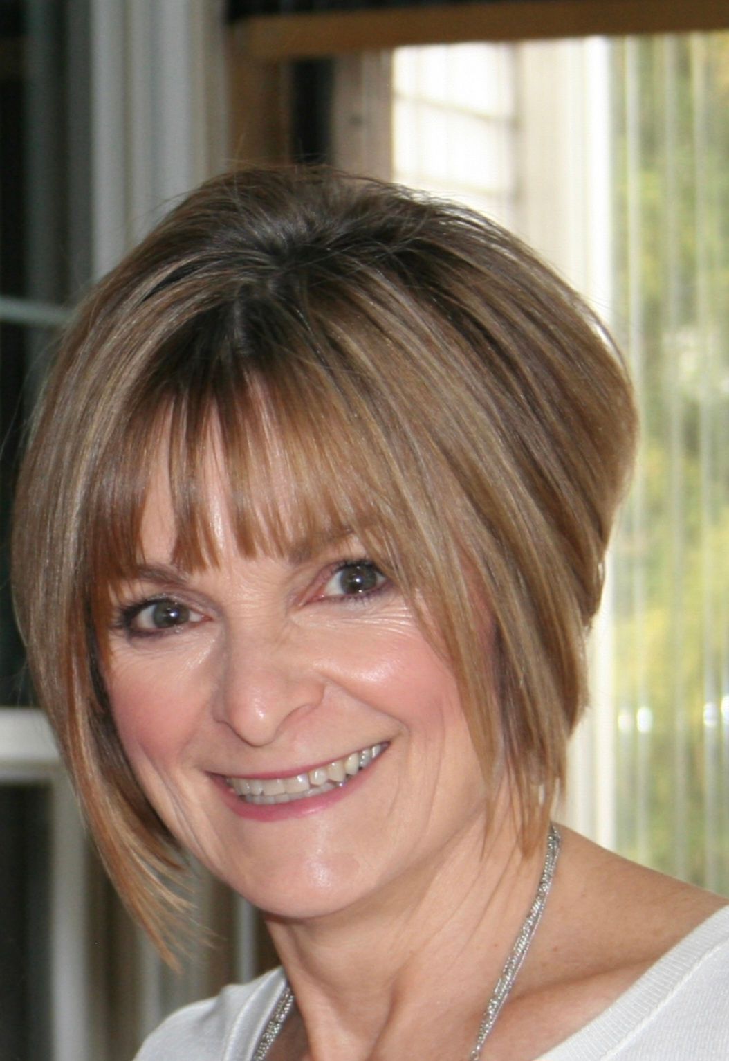 Woman with short, layered blonde hair smiles, wearing a white top, indoors with window in the background.