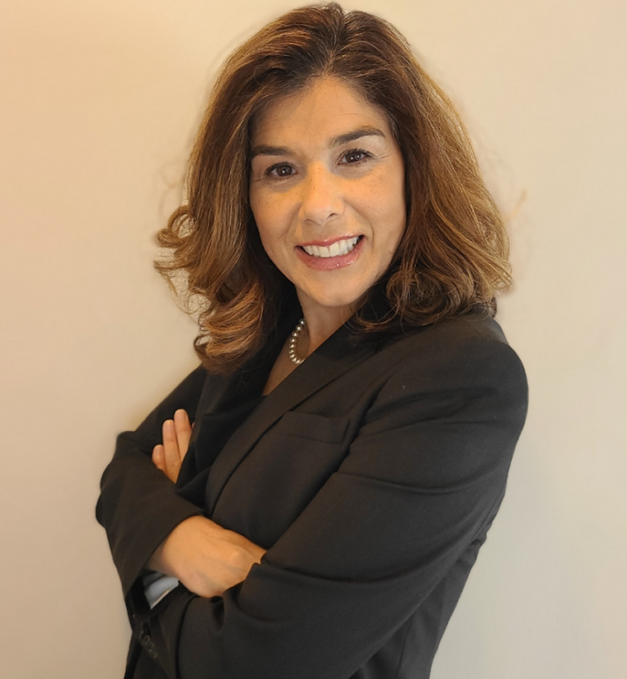 Woman in a black blazer with arms crossed, smiling, posing in front of a white wall.