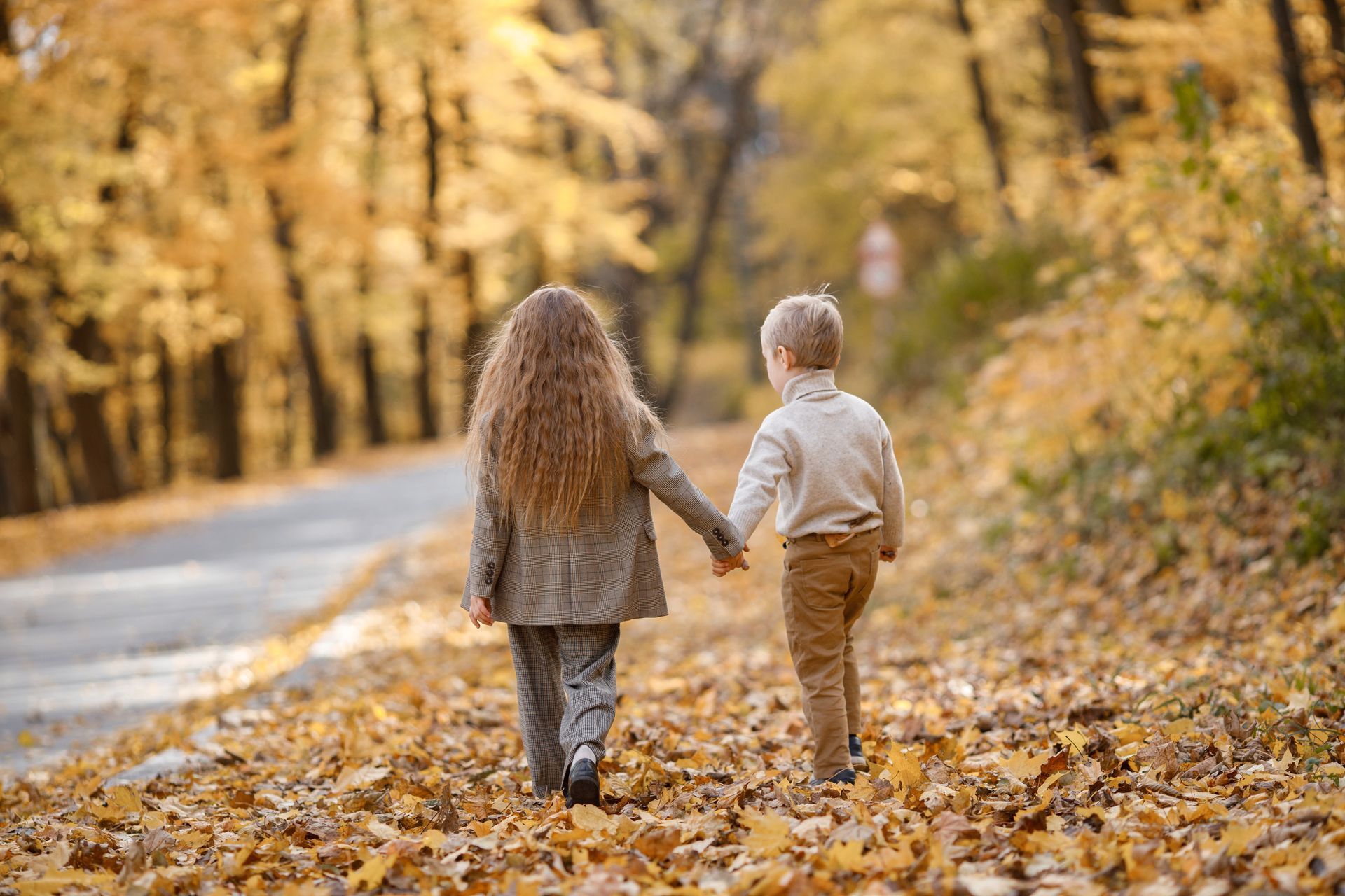 Two children holding hands, walking on a leaf-covered path in an autumn forest.