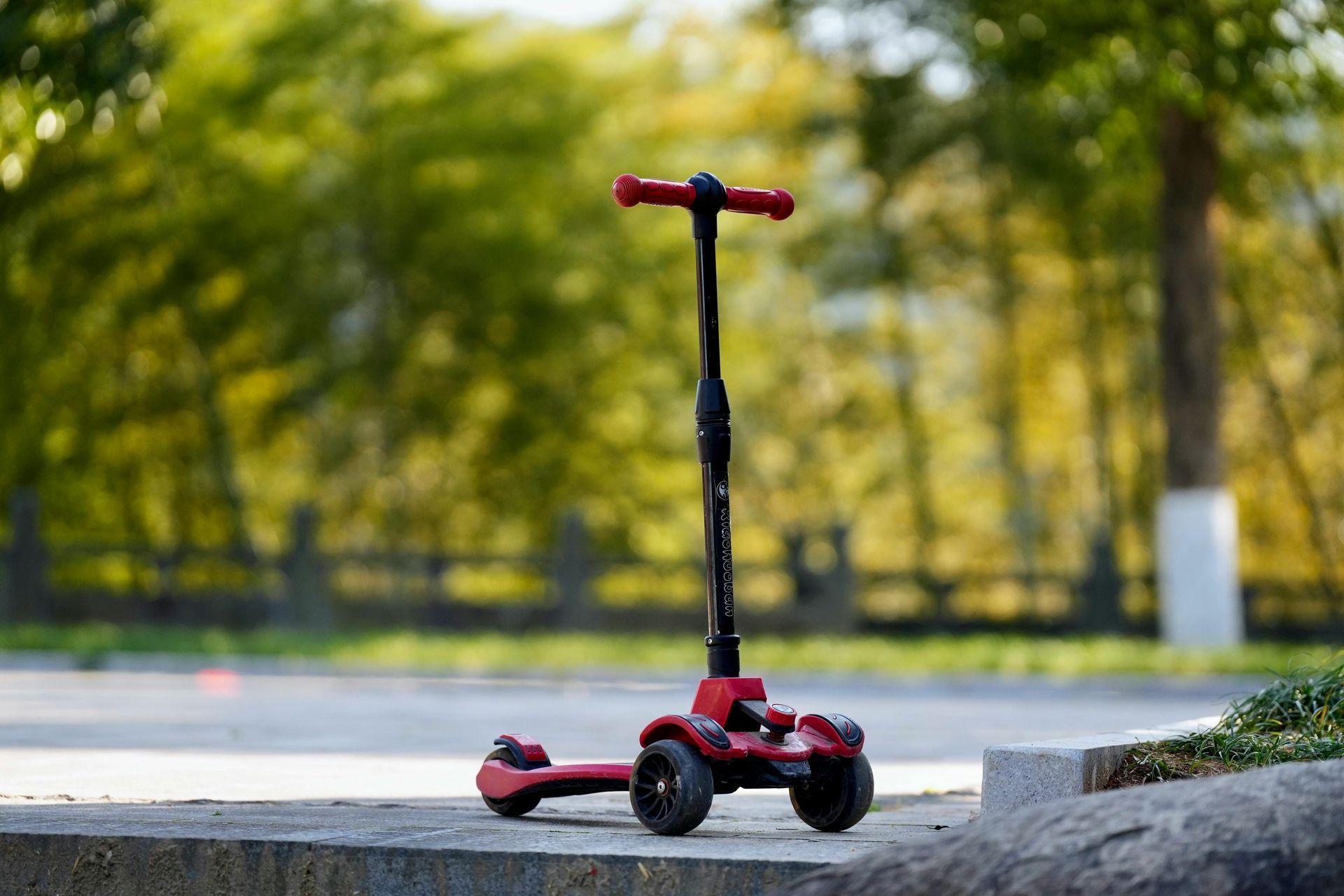 Red and black three-wheeled scooter on a concrete surface, with a blurred green background.