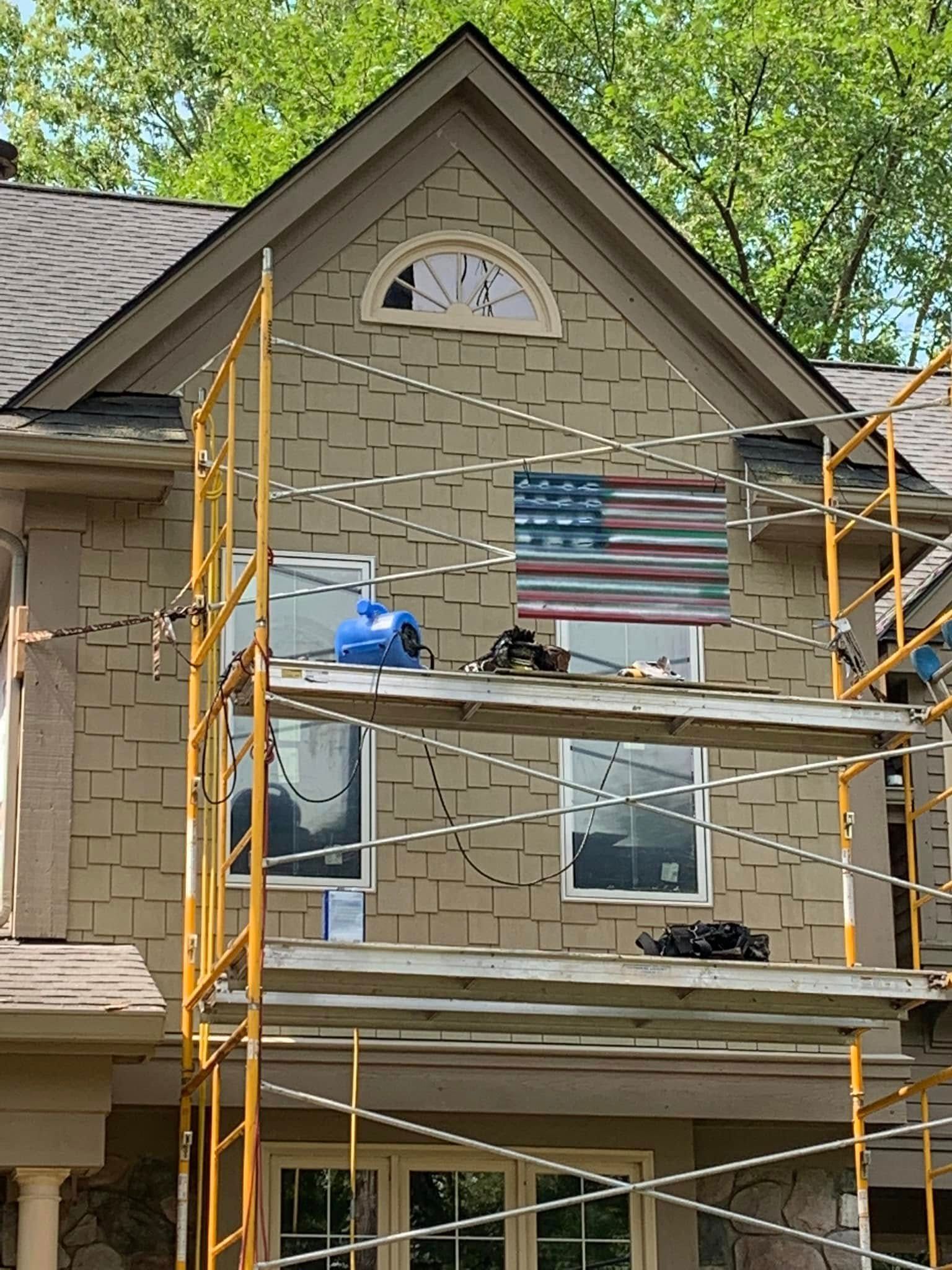 House exterior with scaffolding. American flag artwork. Tan shingles, windows, and a semi-circular window.