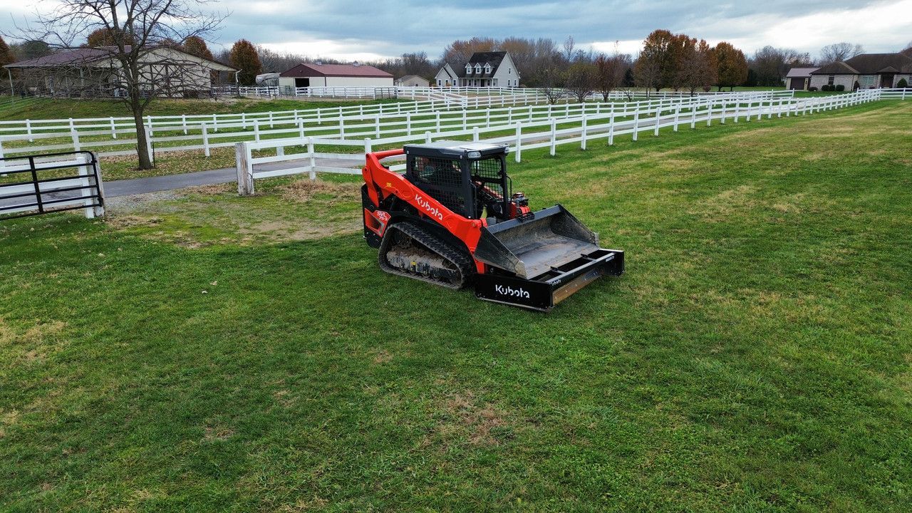 An orange tracked skid steer with a grader box attachment parked on a grassy field near a white fence.