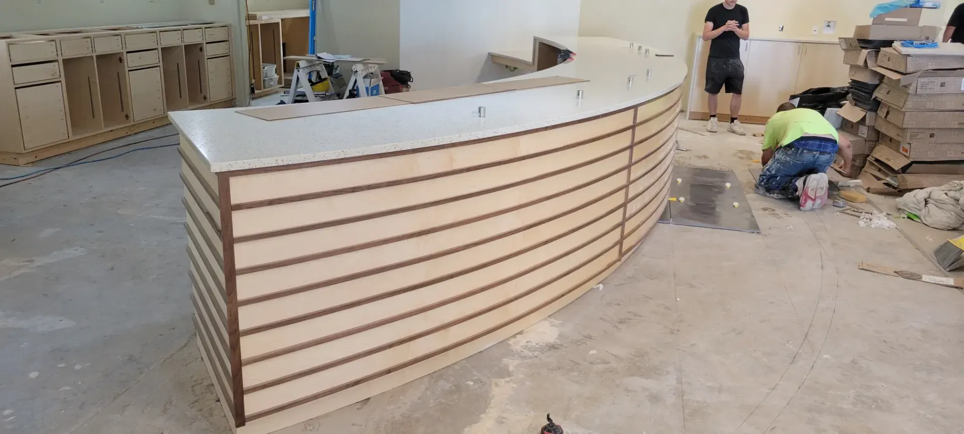 Curved wood reception counter under construction in a workshop, with two workers and tools nearby