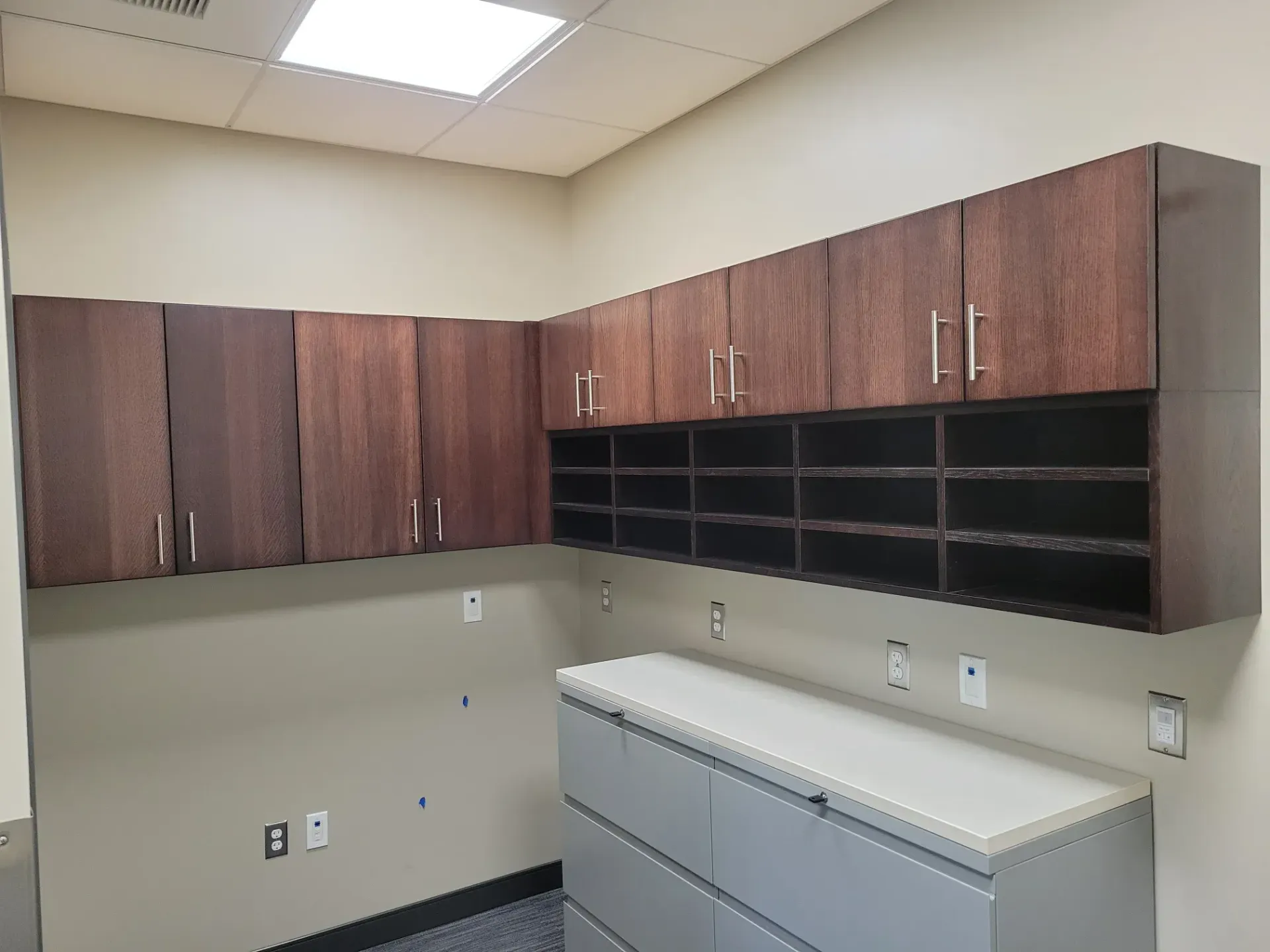 Empty office break room with brown cabinets, black shelving, white counters, and beige walls