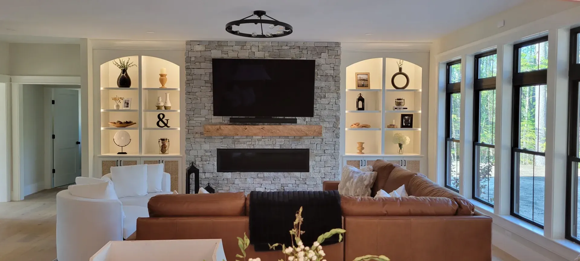 Bright living room with white built-ins, stone fireplace, brown sofa, and large wall-mounted TV