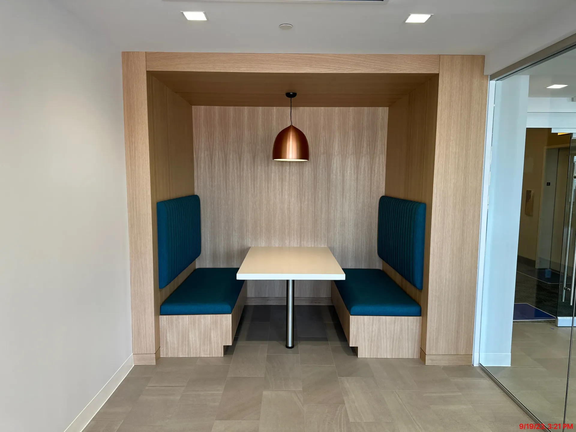 Small booth seating area with teal benches, a white table, wood paneling, and a hanging copper lamp