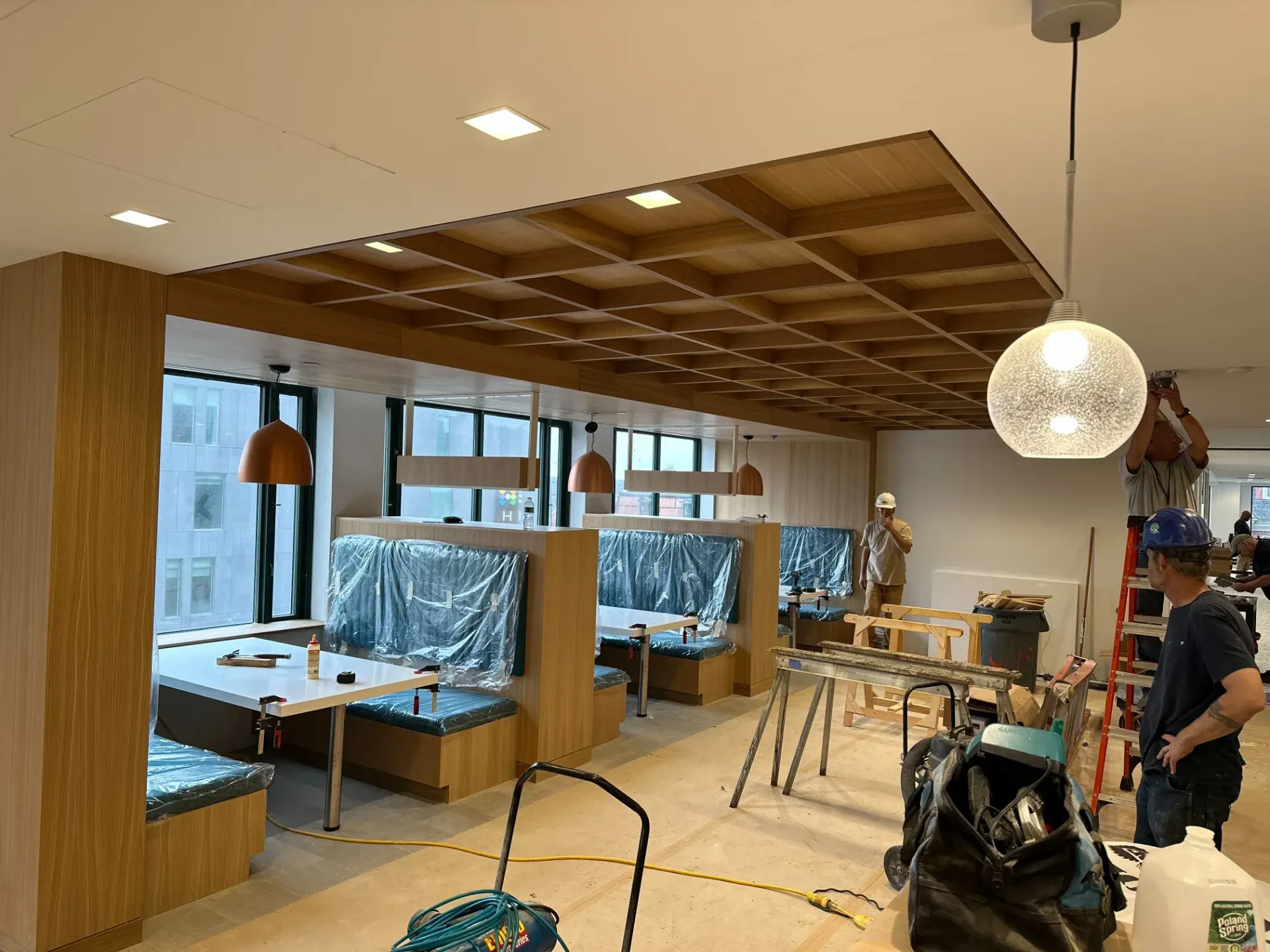 Interior of a workspace with desks, stools, large windows, and a person near a motorcycle under construction.