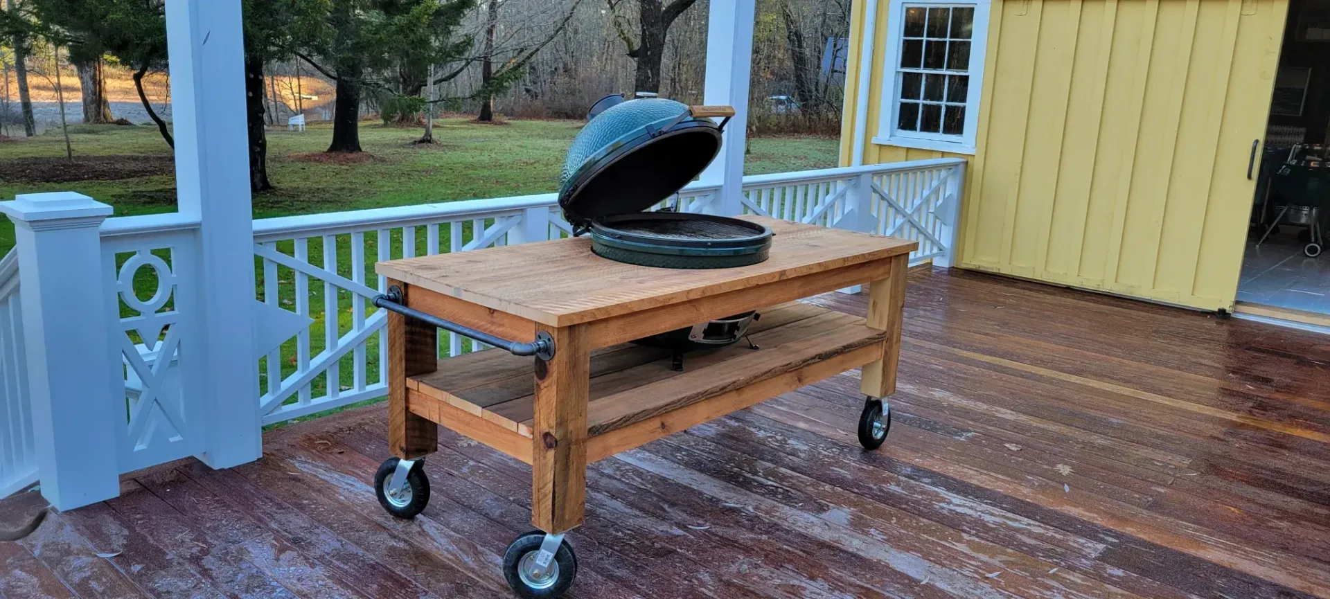 Outdoor wooden table on a wet deck with an open black grill lid beside a yellow house