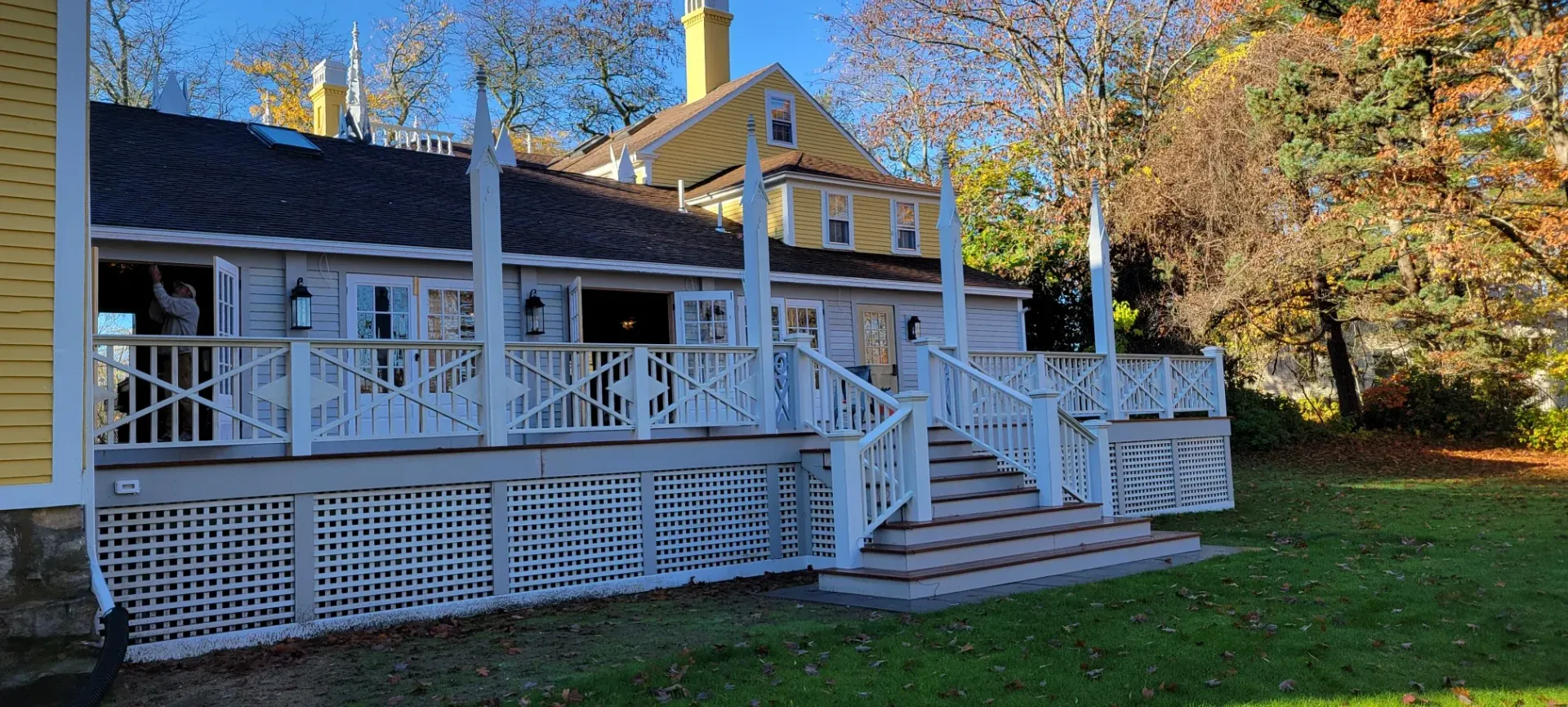 White house with a wraparound porch and steps, set in a grassy yard under autumn trees