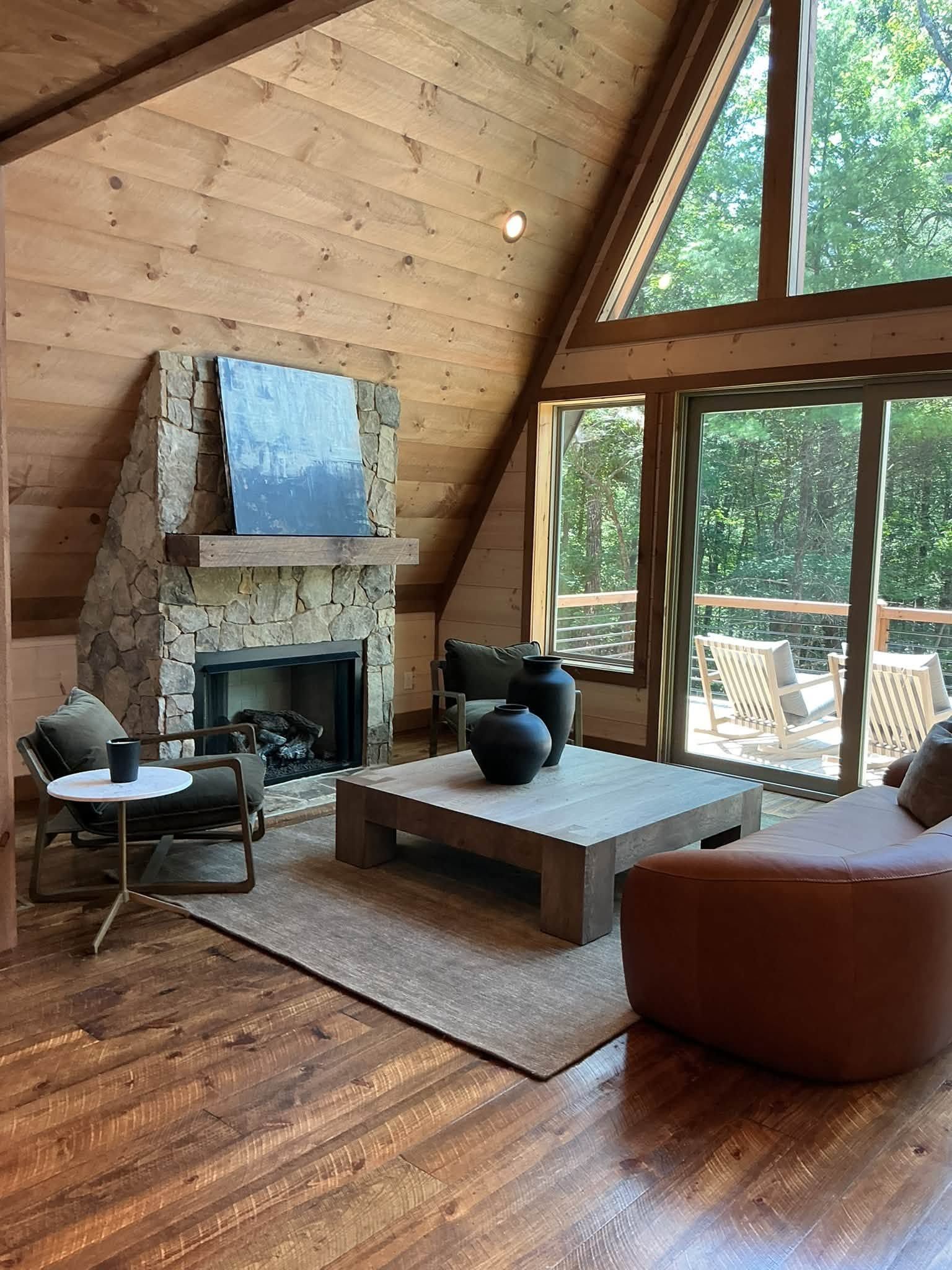 Modern cabin living room with stone fireplace, wood ceiling, large windows, and leather chairs around a coffee table