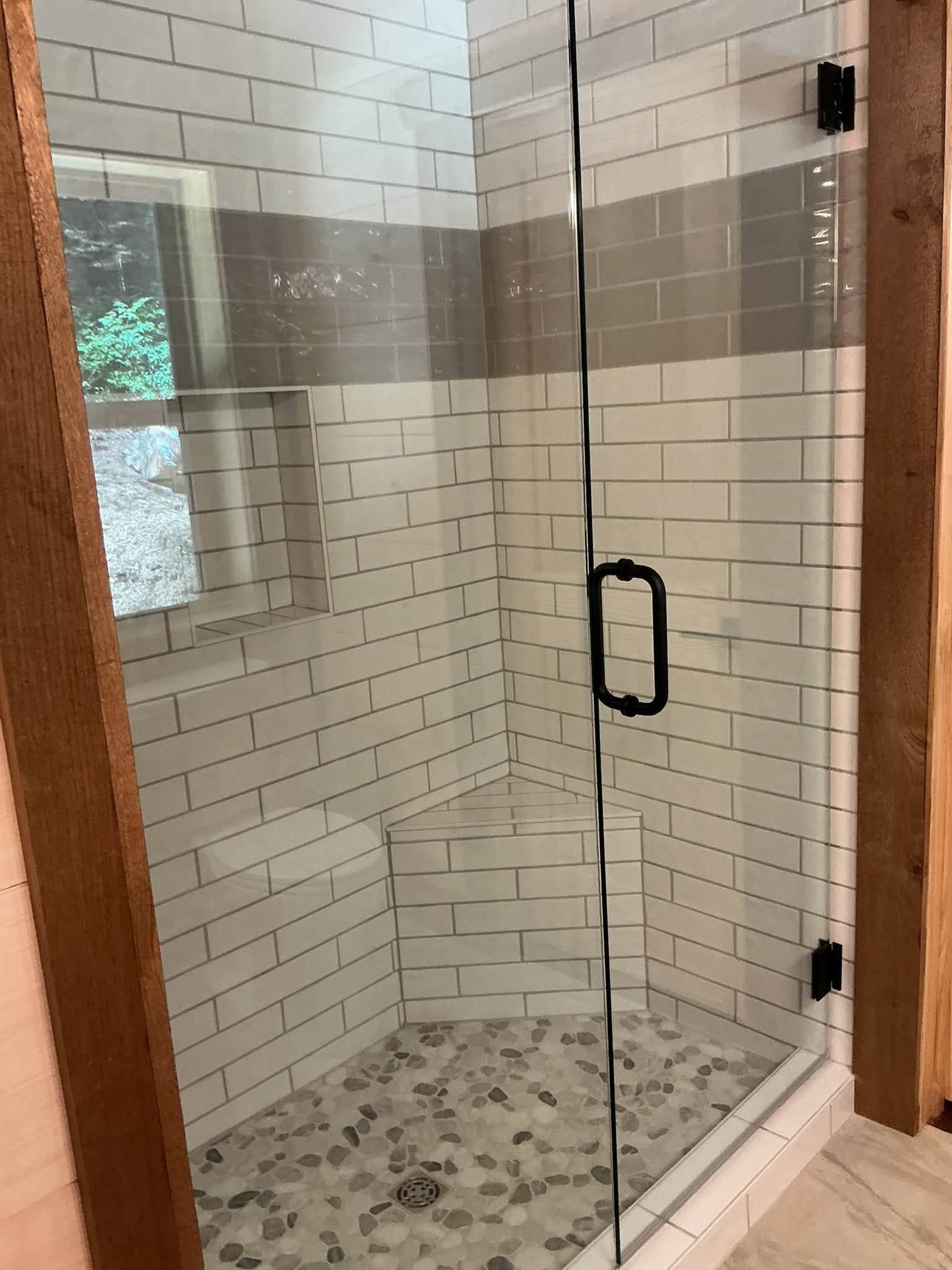 Glass shower enclosure with white tile walls, gray accent band, and pebble floor inside a bathroom.
