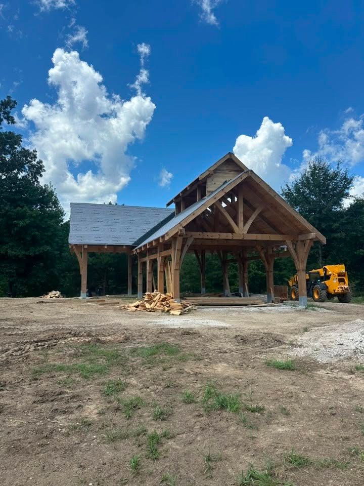 Partially built wooden house frame on a dirt lot under a blue sky, with a yellow excavator nearby