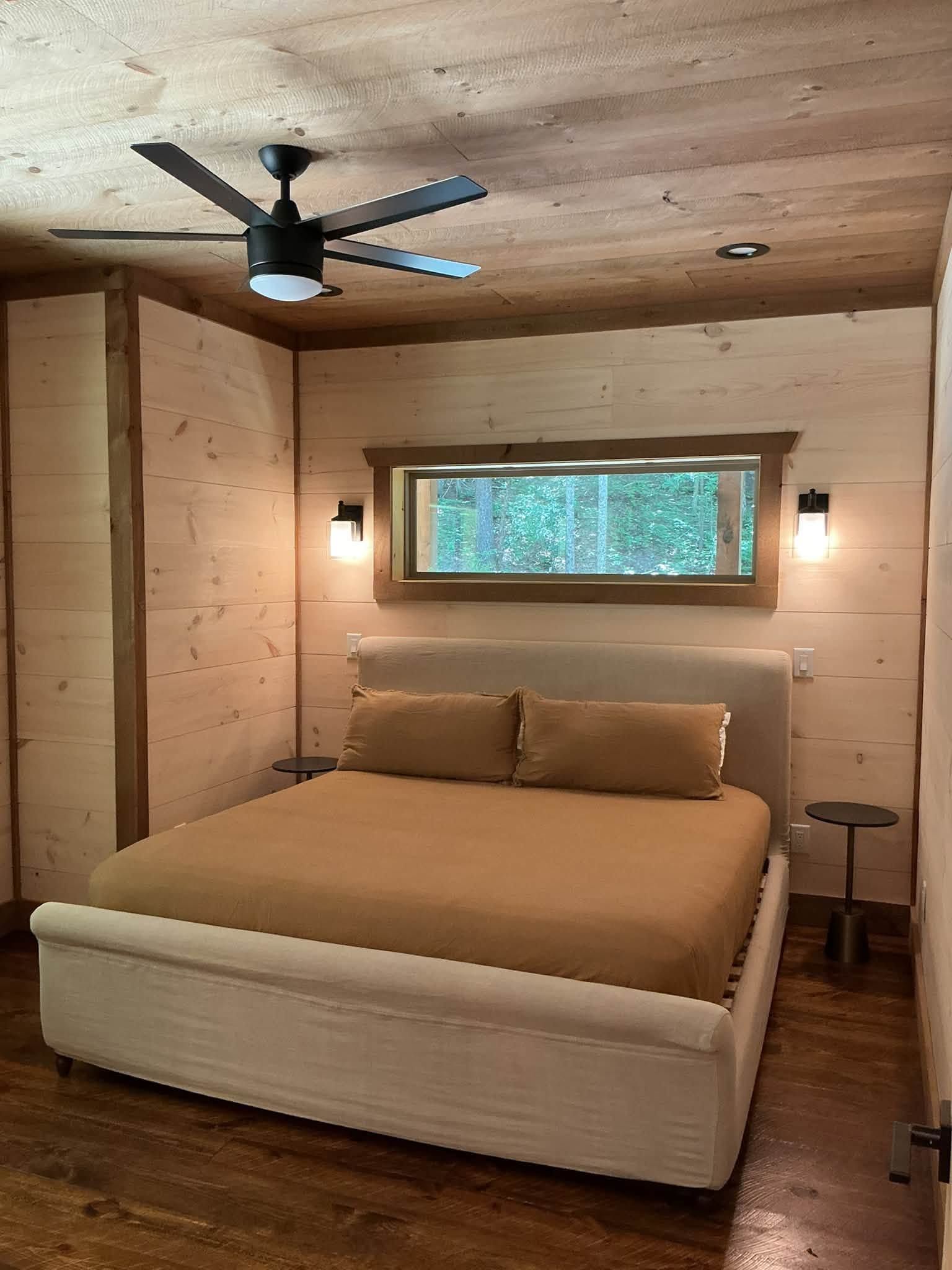 Cozy wood-paneled bedroom with a bed, ceiling fan, wall lights, and a window overlooking trees