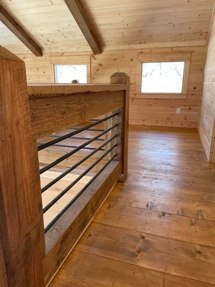Wooden loft interior with exposed beams, railing, and two small windows.