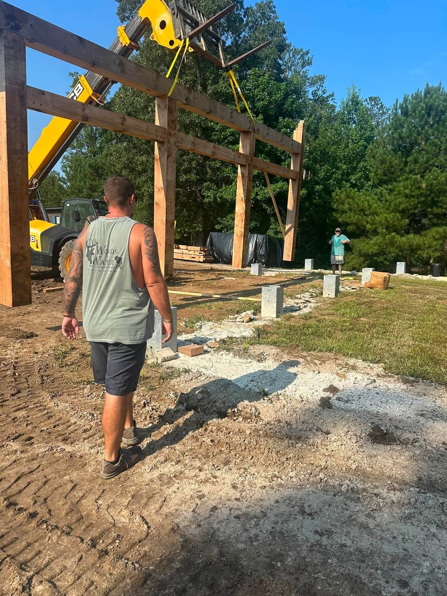 Man walking on a dirt path toward a tall wooden obstacle with a yellow crane overhead under clear blue sky