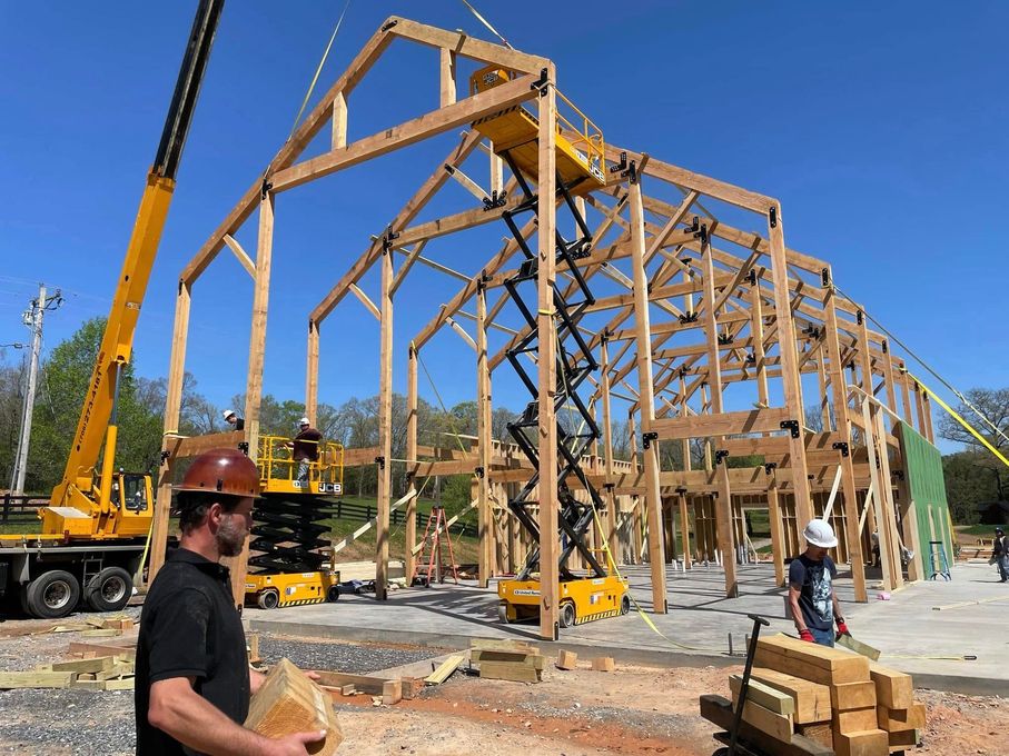 Wooden barn frame under construction with a crane, workers, and stacked lumber on a sunny site