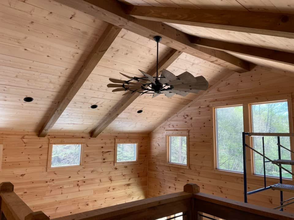 Wood-paneled cabin interior with vaulted ceiling, ceiling fan, small windows, and a loft ladder under construction