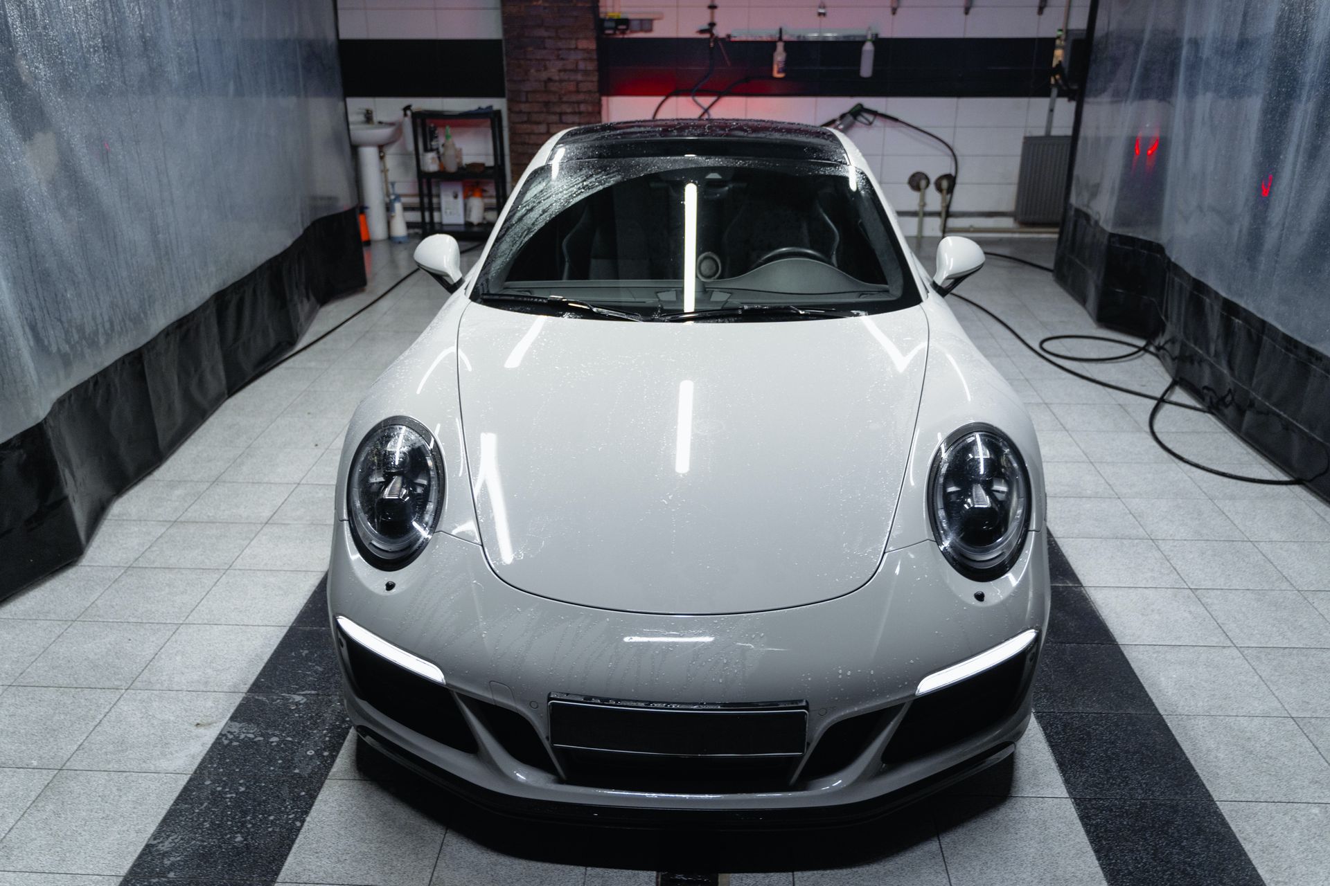 White Porsche 911 being washed in a car wash bay, with wet car and headlights on.