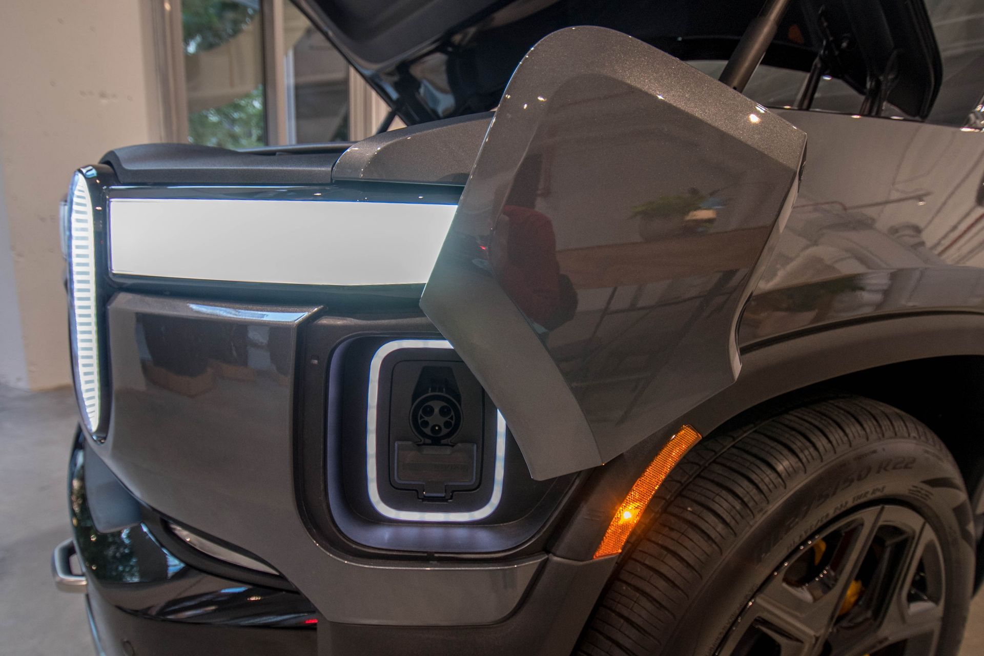 Close-up of a Rivian electric vehicle's front. Open charging port, gray paint, amber side marker, and black tire.
