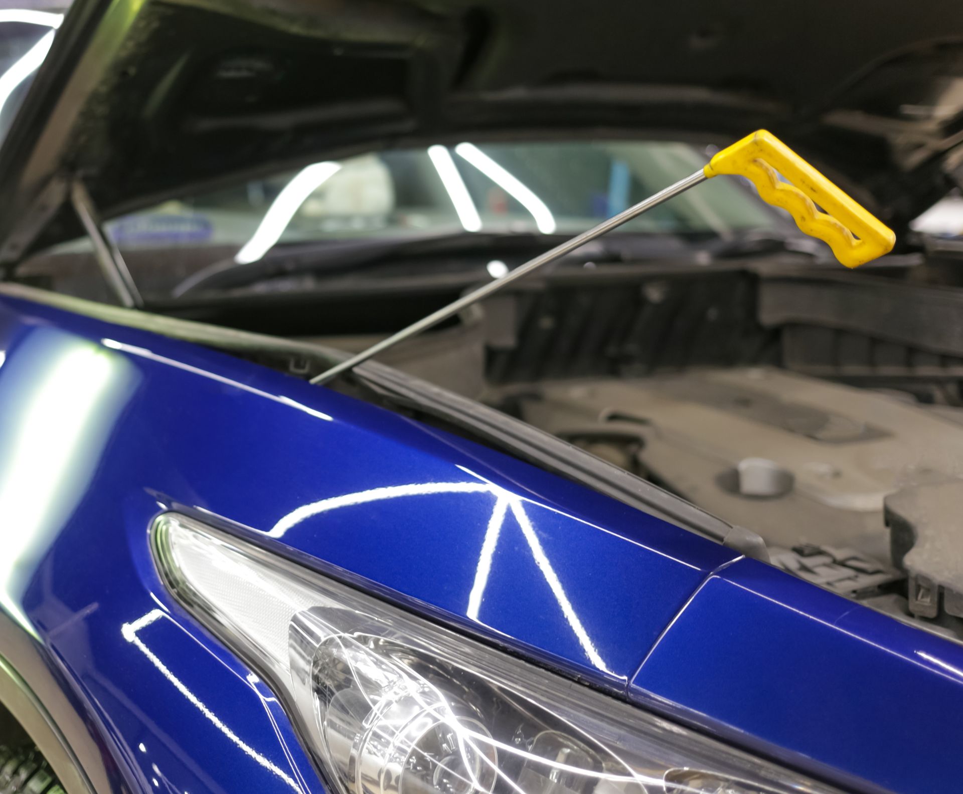 A dent puller tool being used on a blue car's hood in a workshop. Yellow handle, open hood.