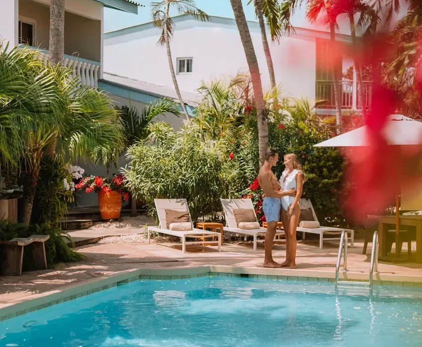 Couple standing by a pool in tropical setting, embracing. Lush greenery and white buildings in background.