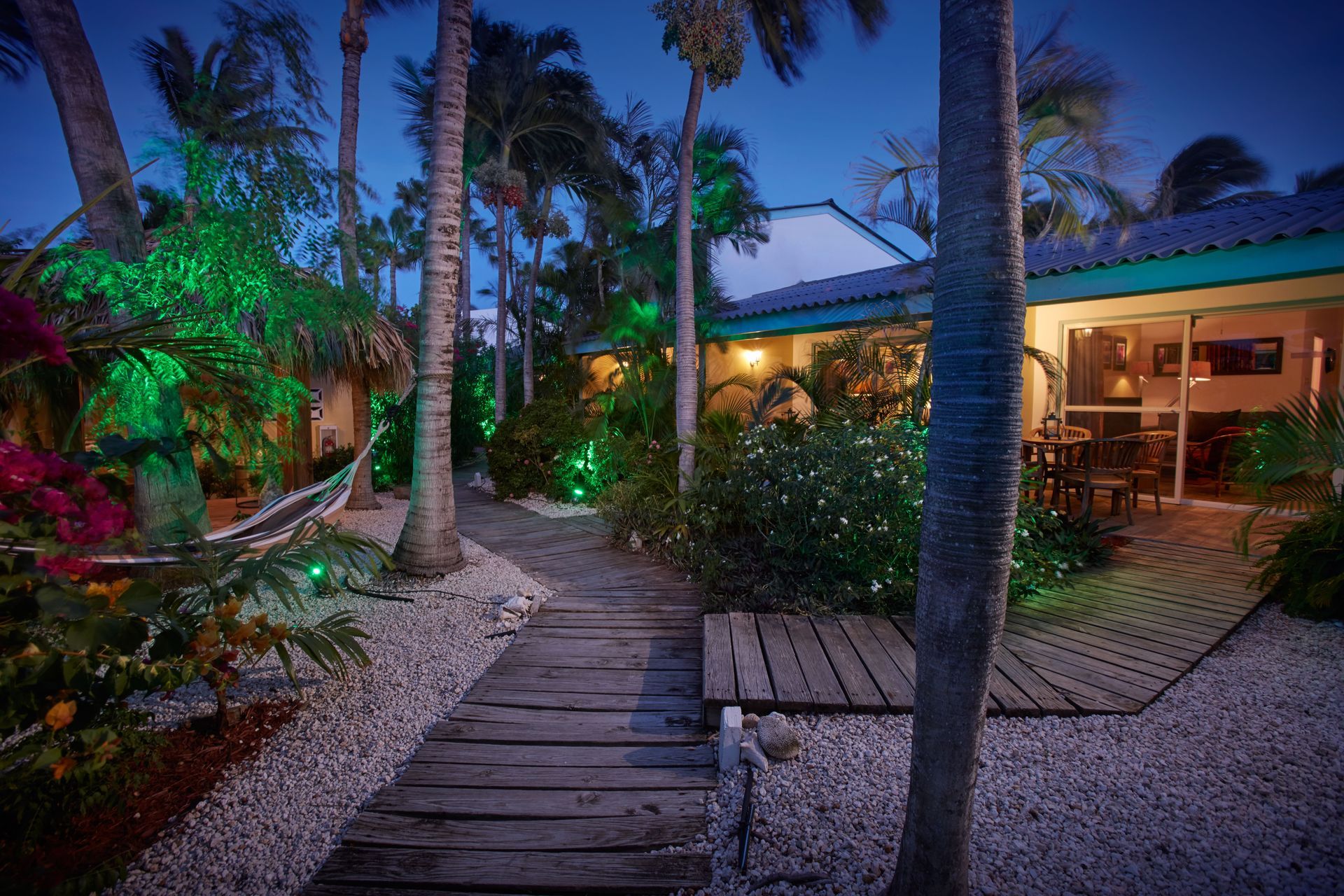 Night view of a tropical resort with a wooden walkway, palm trees, and illuminated green lights.