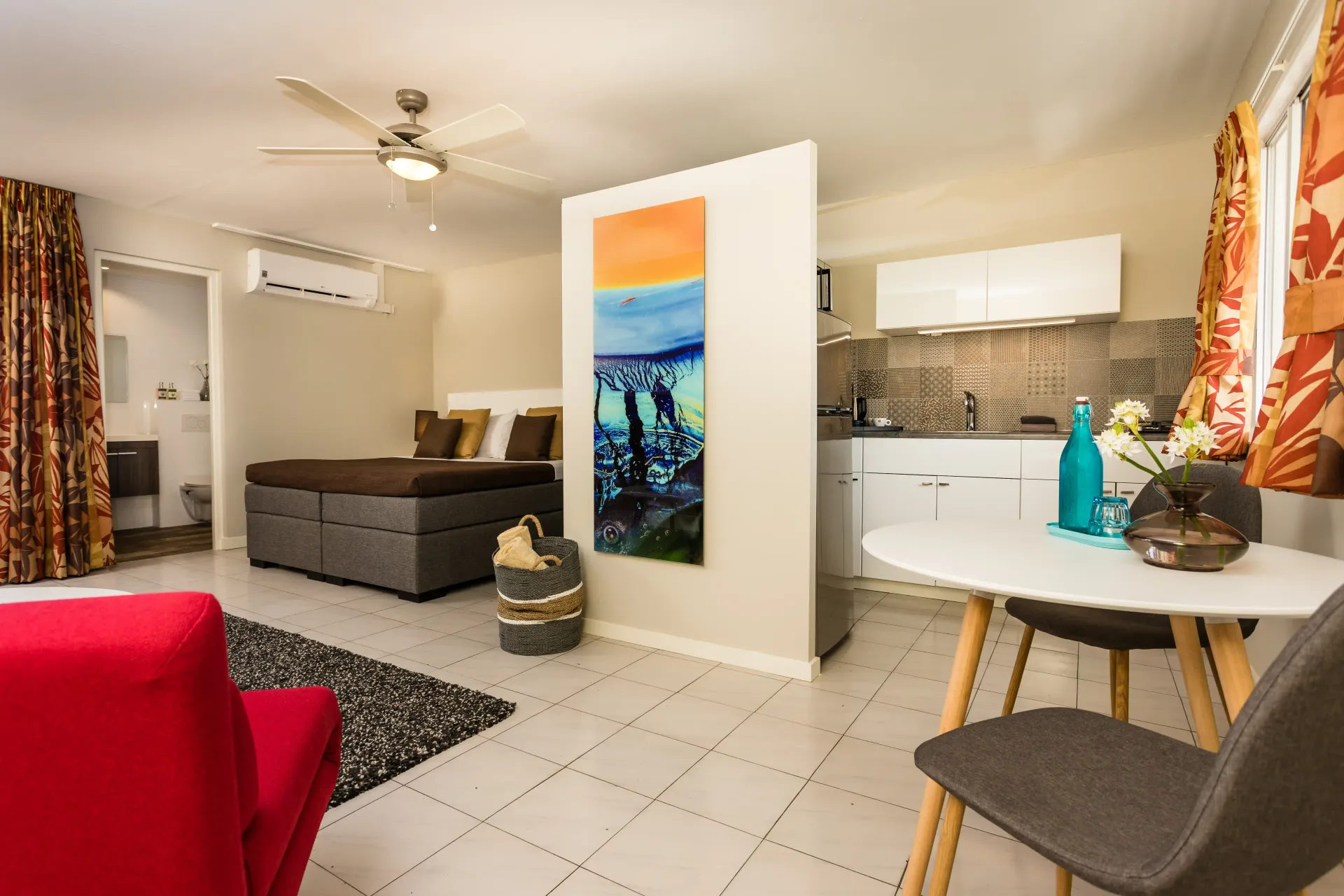 Studio apartment with red armchair, kitchen, bed, and artwork; white tile floor.