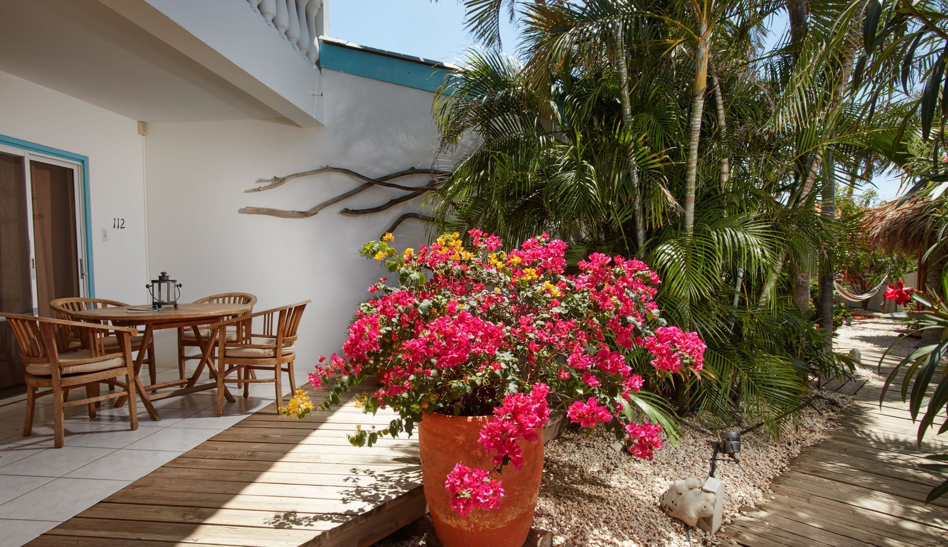 Patio with wooden furniture, flowering plants, and white building with blue trim.
