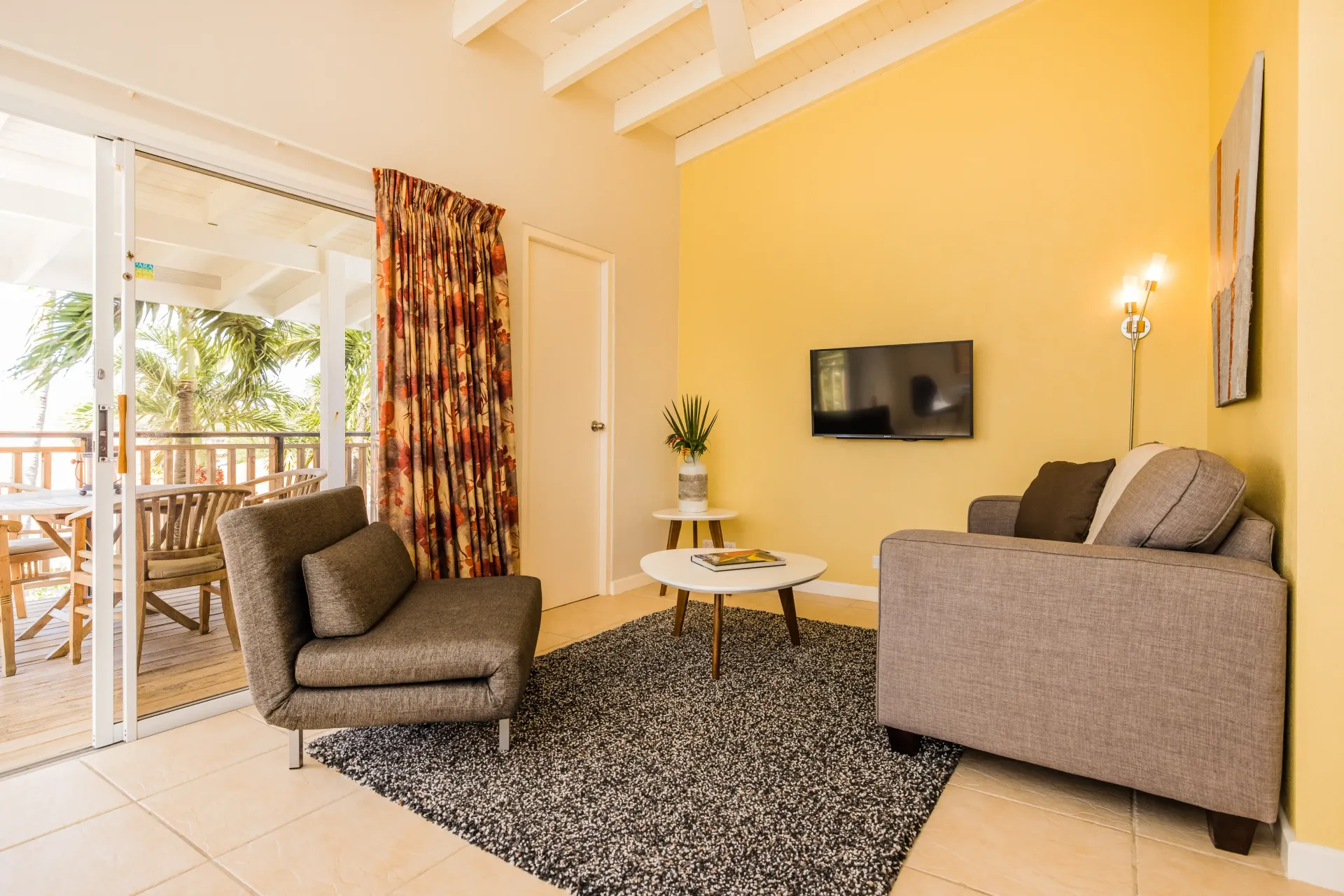 Living room with open doors to a balcony, yellow walls, gray rug, two sofas, and a TV.