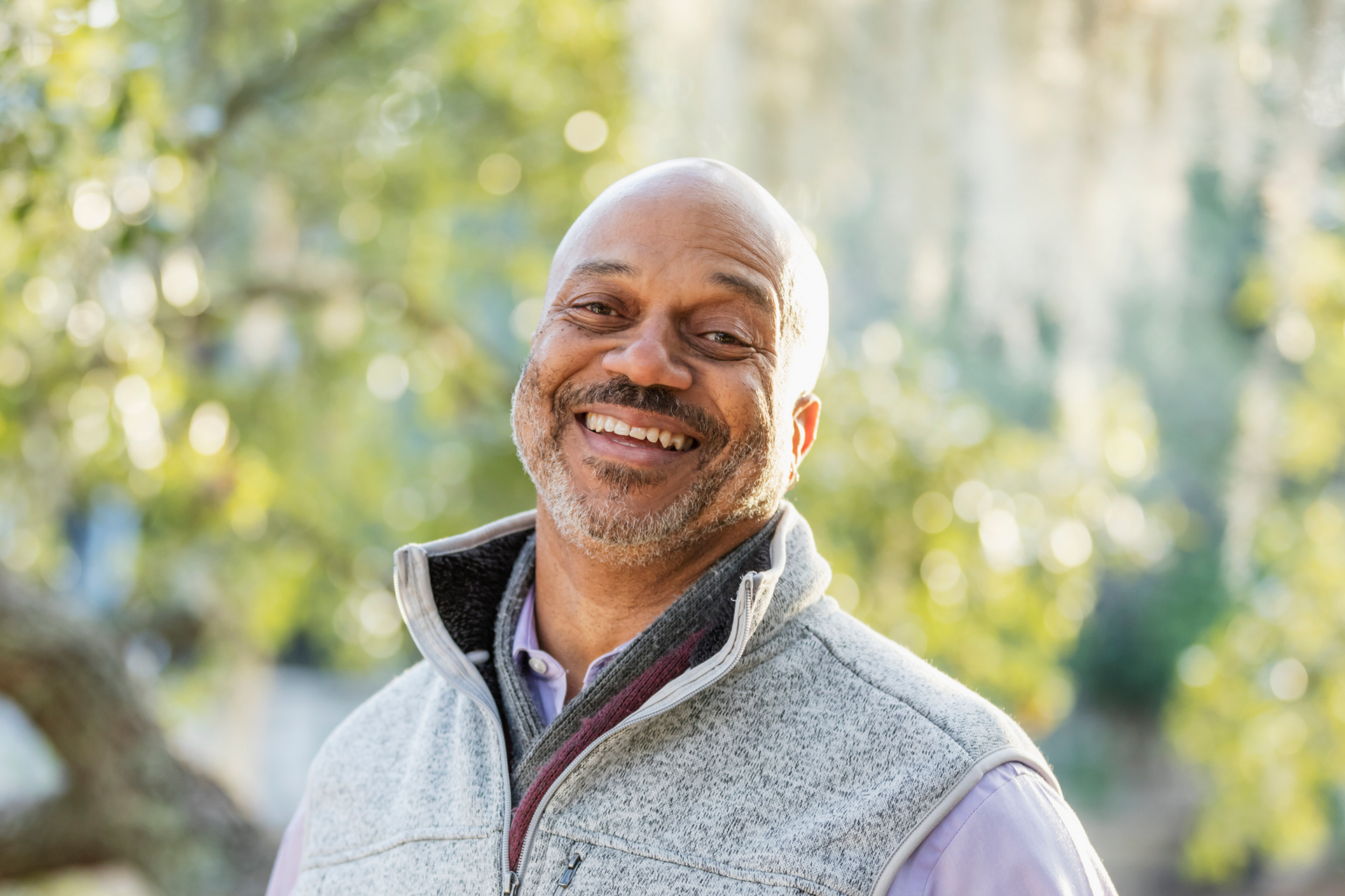 Smiling man in a light gray vest outdoors with sunlit greenery in the background