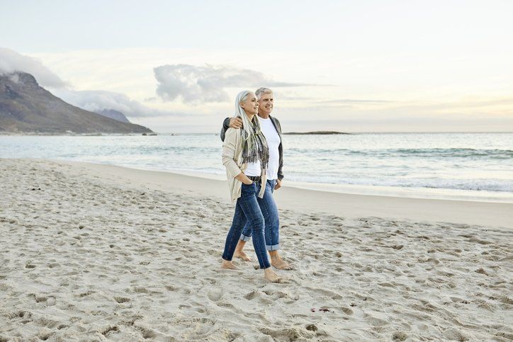 Two people walking on a sandy beach near the ocean at sunrise, wearing jackets and jeans
