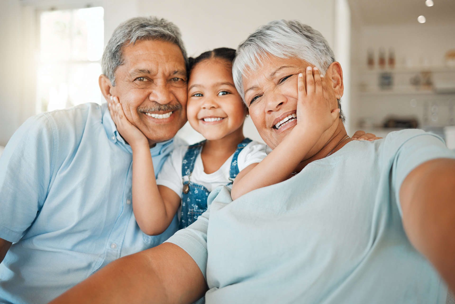 Three smiling family members sitting close together in a bright home, wearing light blue tops.